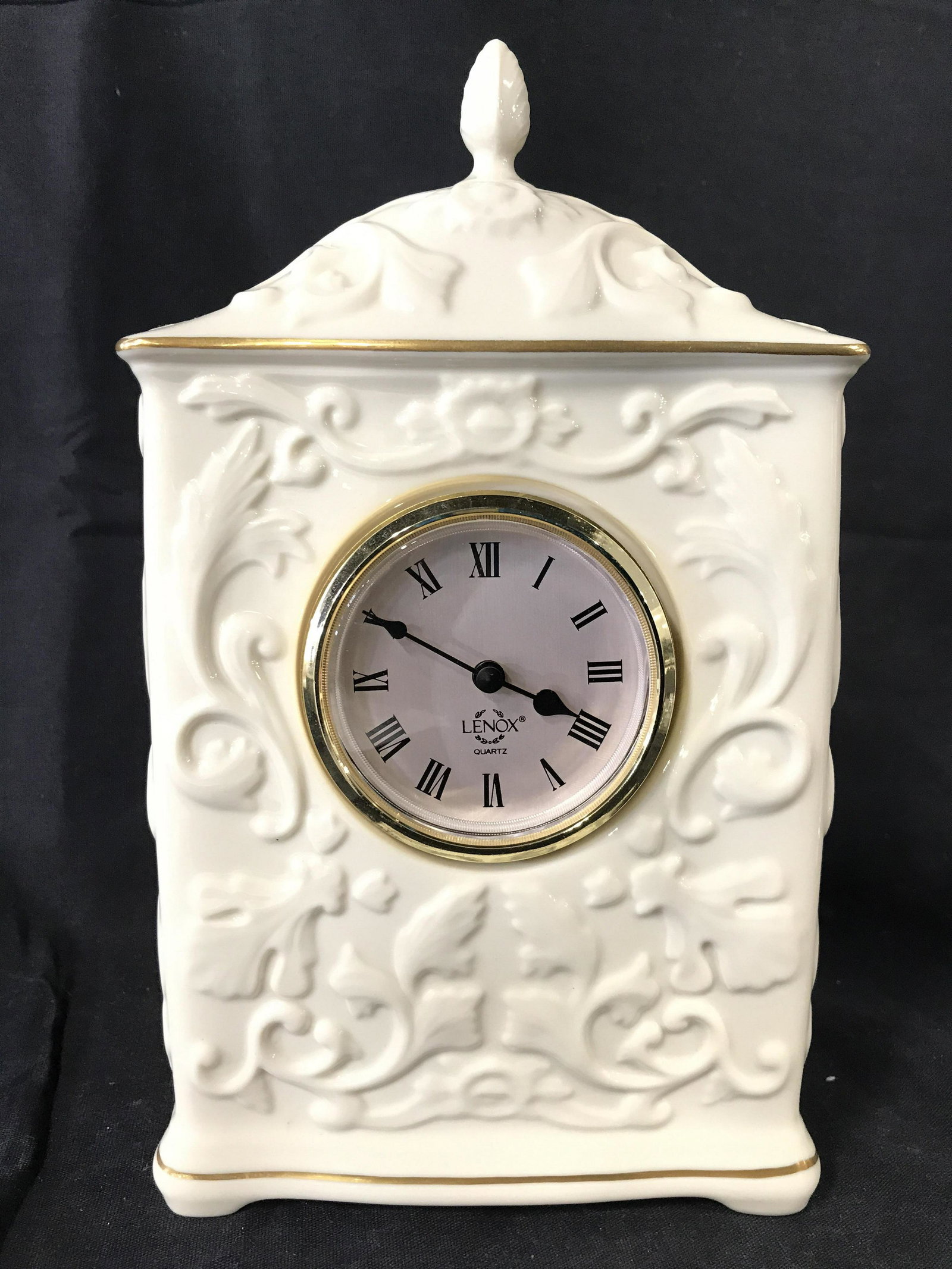 LENOX Porcelain Desk Clock: Clock face is marked LENOX. Clock face is silver toned with black toned numericals. Piece is porcelain and cream toned with gold toned accents. Top of piece has finial style structure. Lenox clock, Le