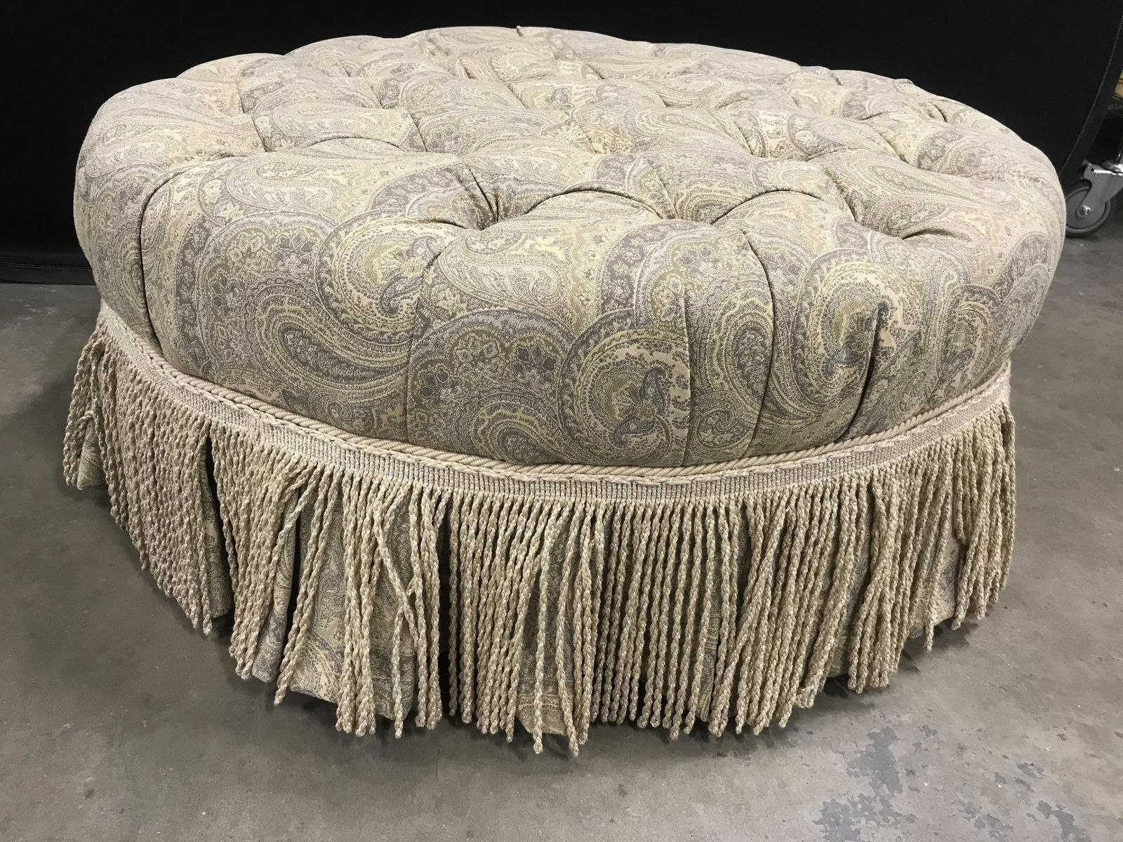 Ethan Allen Upholstered Tufted Ottoman in Casters: Good condition, Ethan Allen Button Tufted ottoman in neutral paisley fabric with bullion fringe, seat, bench, hassock, property of Wilton Connecticut estate, measures ht 16 inches diameter 37 inches,