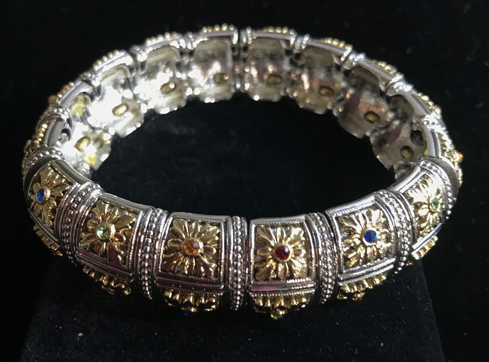 Judith Ripka Style Crystal & Bi Color Tn Bracelet: Embossed Metal panels with 2 gold tone flowers, center colored Solitaire crystal and other designs in relief. Wired on a fish wire. app 3 in. diameter. Slightly weighted piece. Property of Upper East
