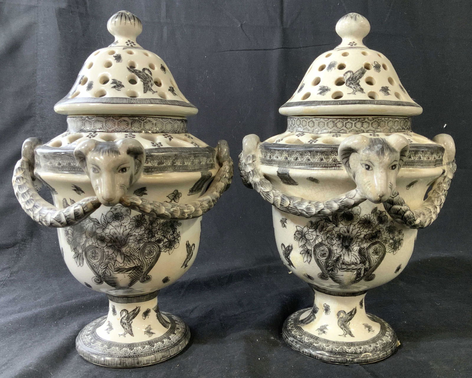 Pair Hand Painted Signed Rams Head Urns: Pair signed Ceramic urns, lidded urns, open lattice detailing, swags stand proud to vessel, each has two rams heads as handles, each has pedestal base, each signed underside illegibly, each marked for