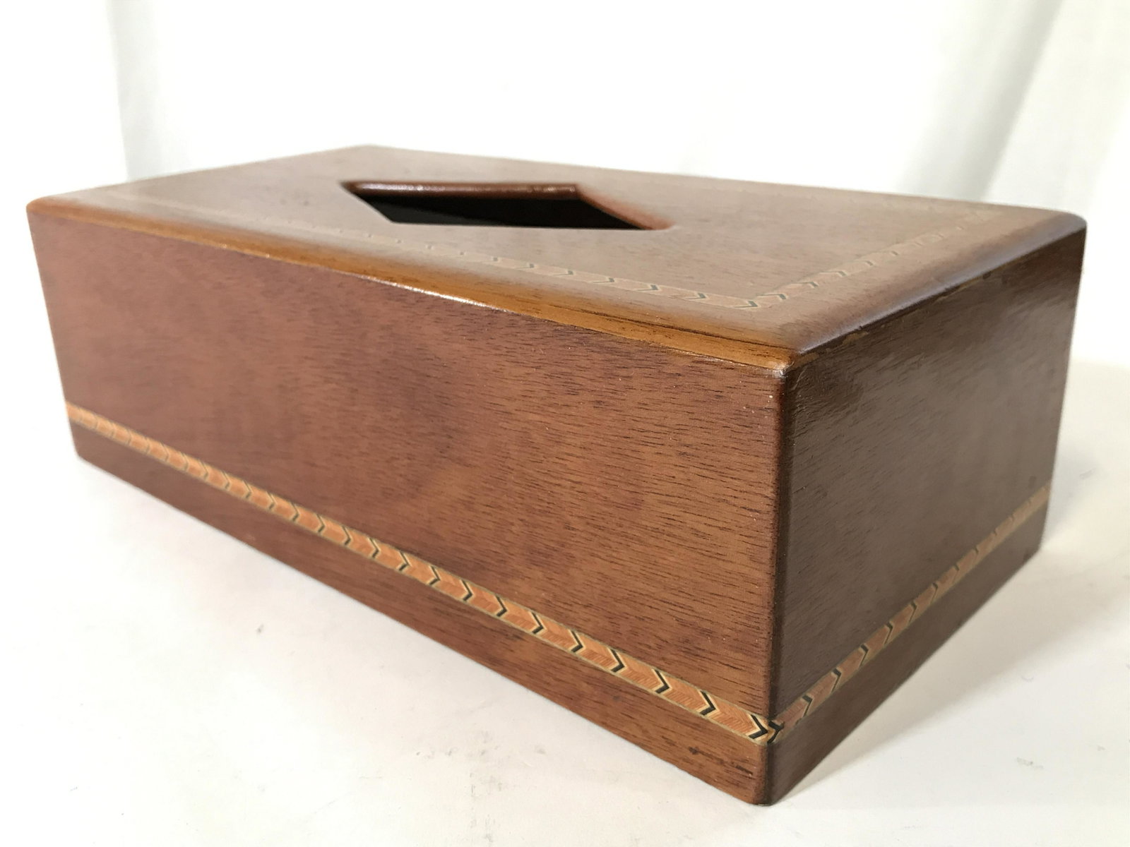 Inlaid Wooden Tissue Box Holder: Tissue box holder is wooden. Piece appears to have inlaid detail with black and gold toned arrowhead shape pattern detail. Piece has diamond shaped opening. Piece measures approximately 11 inches long