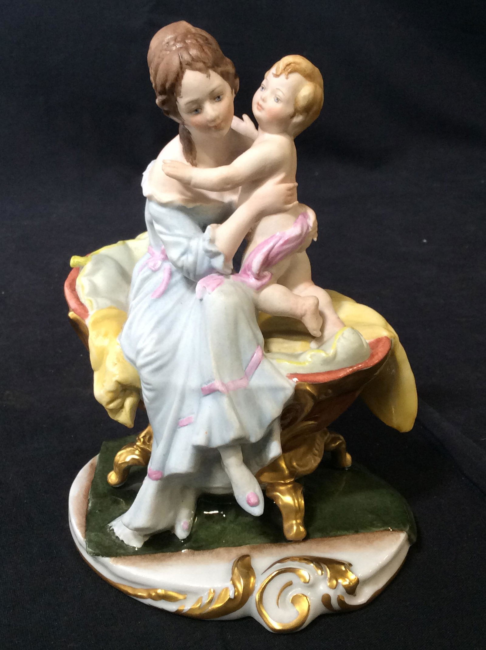 Capodimonte Woman W Child Figural, Signed B Merli: Signed Fine Porcelain Of Woman sitting on child’s bassinet holding child. She is dressed in her long light blue gown, Gild accents, Hand painted, underside marked in blue ink, crown over N, reads Ki