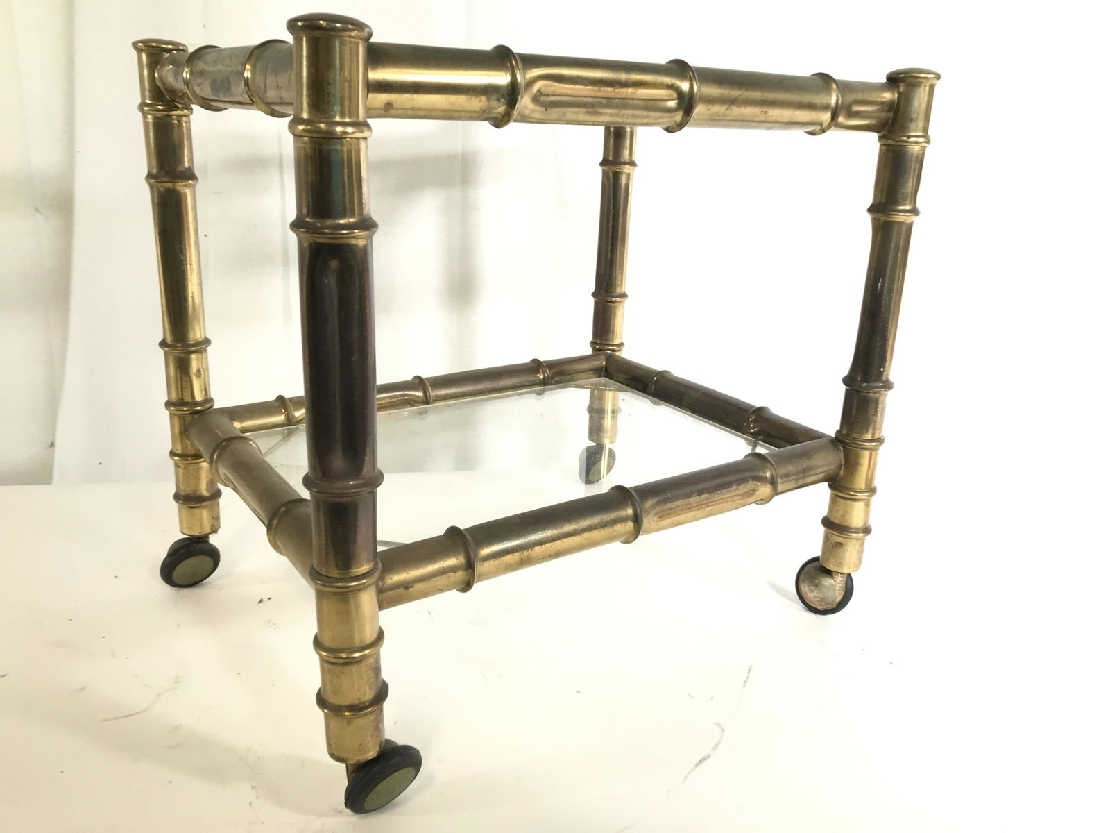Vintage Brass Side Table W Glass Top On Casters: Thick faux bamboo style brass frane, glass table on casters. Side table is gold toned and brass. Piece is vintage possibly antique. Table has glass top and lower glass level. Piece sits on 4 casters.