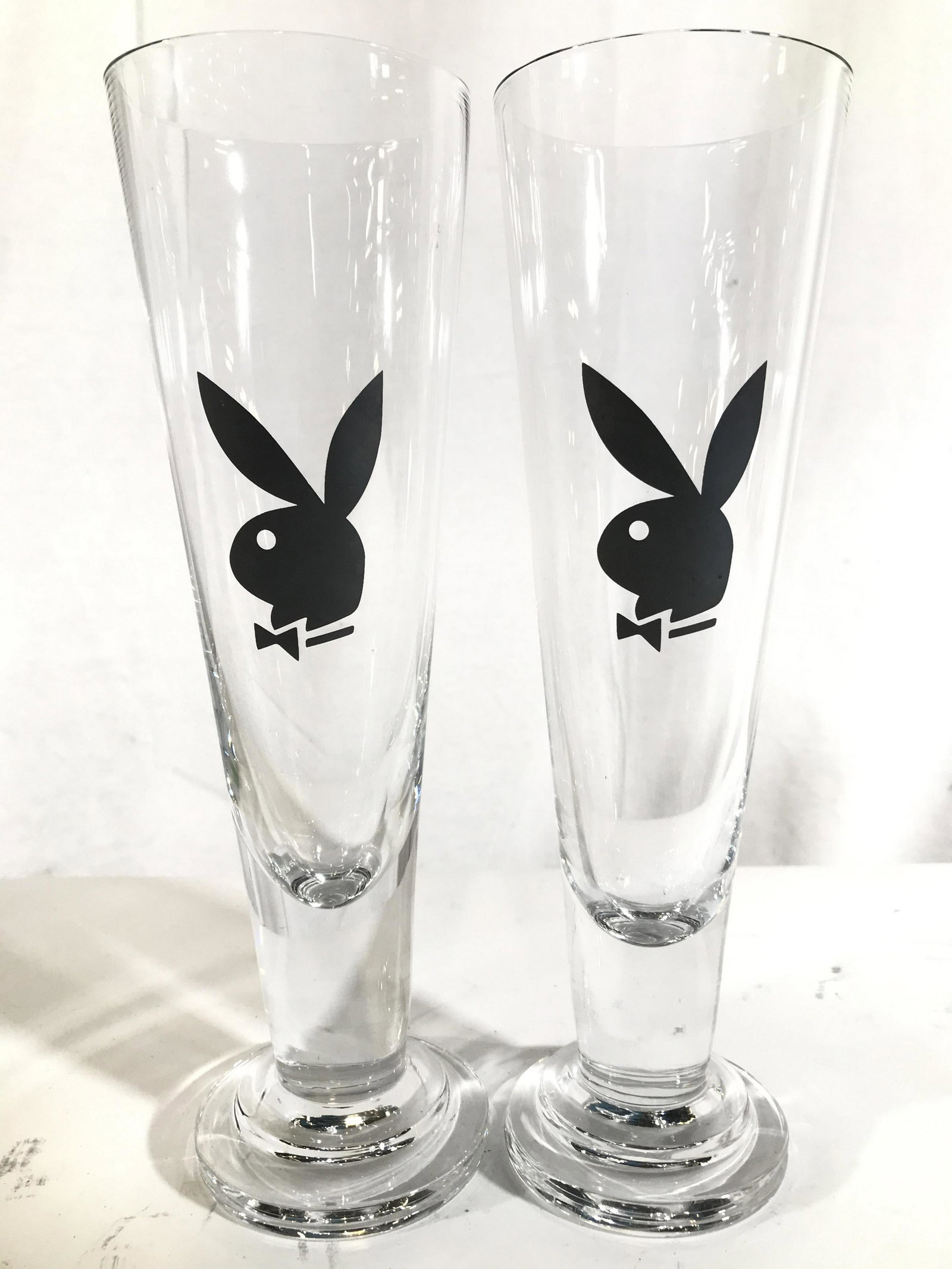 Pair Playboy Bunny Champagne Flutes: Lot includes 2 champagne flutes. Each have a black toned playboy bunny logo on front. Each piece measures approximately 10 inches tall 3 inches wide. Champagne flutes, playboy champagne flutes, playbo