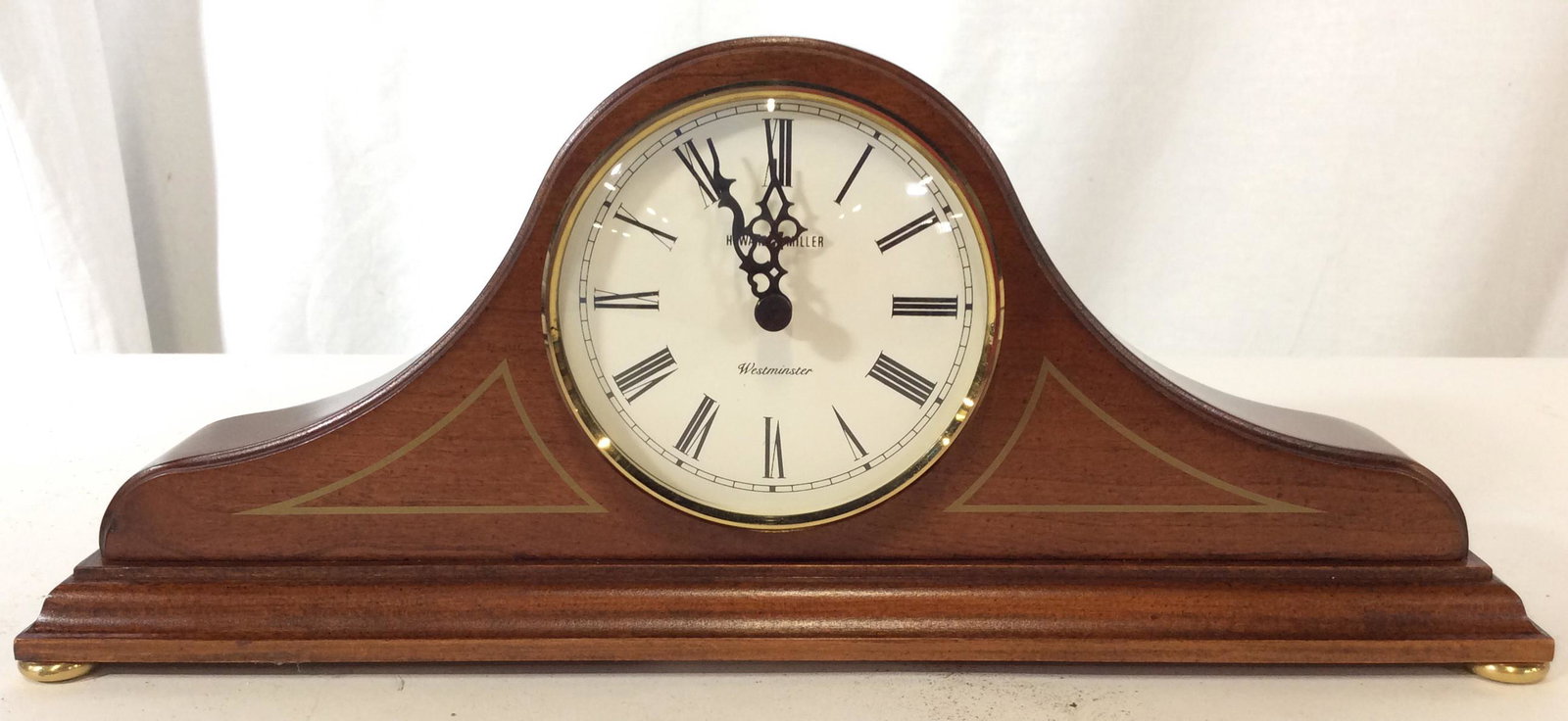HOWARD MILLER WESTMINSTER Mantel Clock: Clock face reads HOWARD MILLER WESTMINSTER. CLOCK is wooden. Clock face is cream toned with black toned Roman numerals and black toned hands. Clock has arched shape and gold toned feet. Piece measures