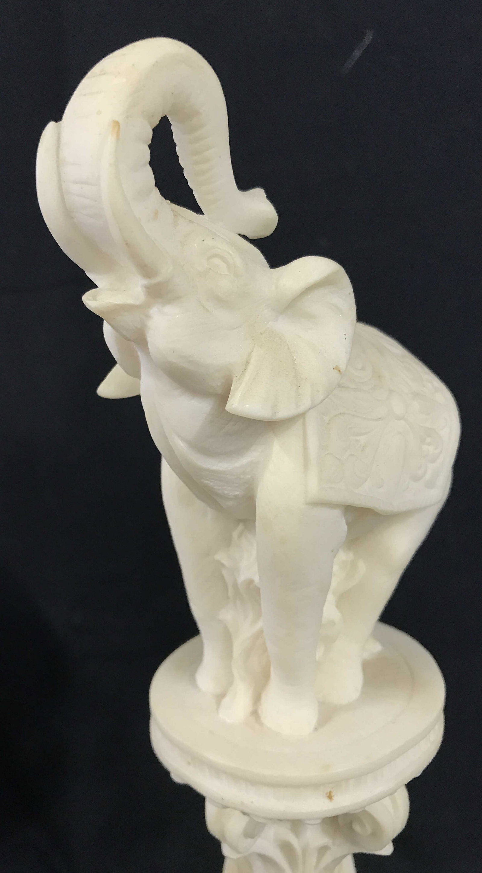 Carved Elephant Pedestal Sculpture: Measures approx 13.5 inches H, and 3.75 inches W. Piece is hand carved and appears to be crafted from a composite material. Elephant sculpture, mantle accessory, elephant themed sculpture, pedestal, d