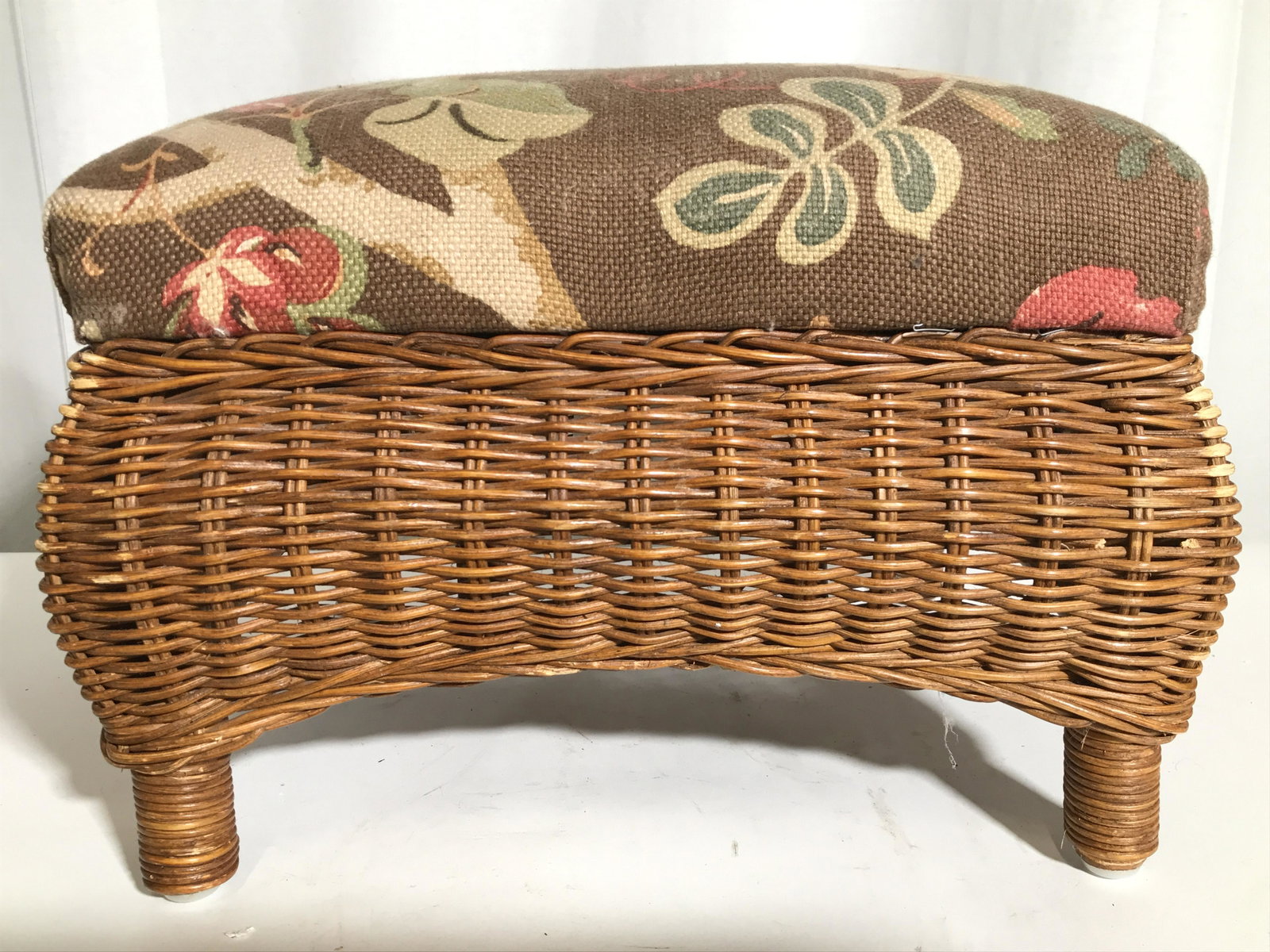 KIRKWOOD INDUSTRIES Upholstered Wicker Footstool: Underside has tag which reads KIRKWOOD INDUSTRIES. Footstool has upholstered cushion with multi toned floral and leaf detail. Piece has a dark beige tone with woven wicker structure. Piece measures ap