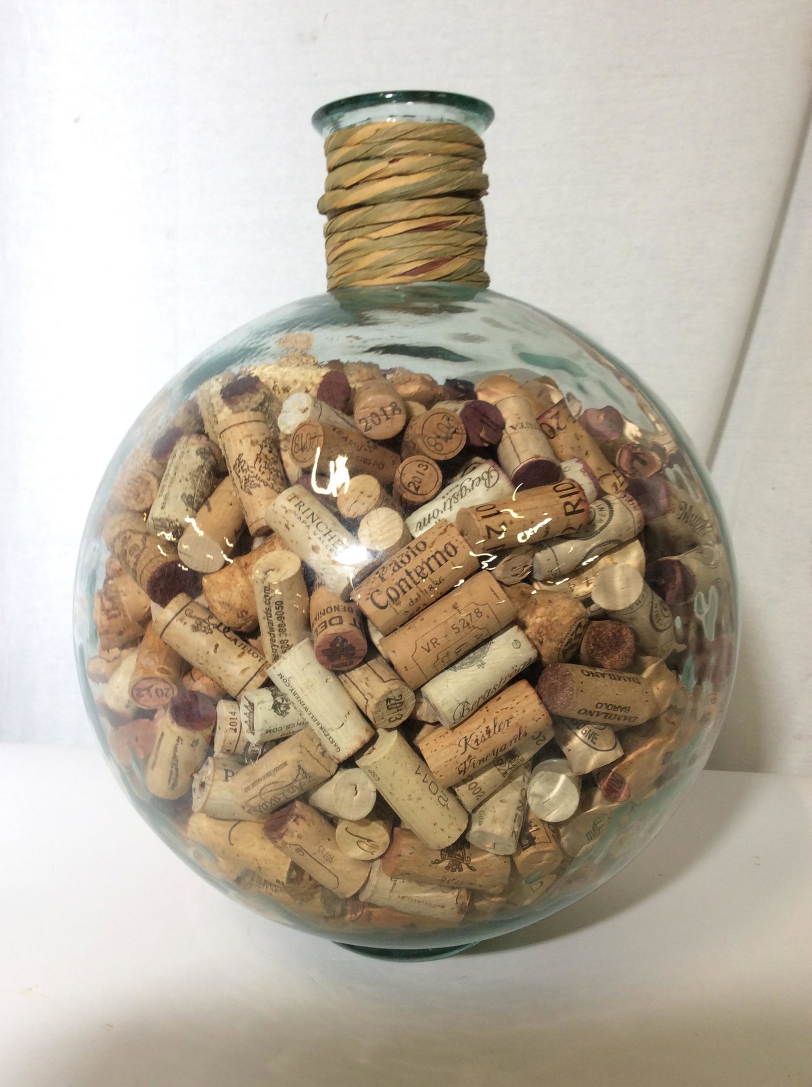 Centerpiece Glass Decor Vessel with Wine Corks: A compliment to any table, counter, kitchen, display. Thick Walled round glass jar, slight blue green hue to glass, wrapped ruffian around bottle neck, numerous corks to count. Open top, no stopper. 1