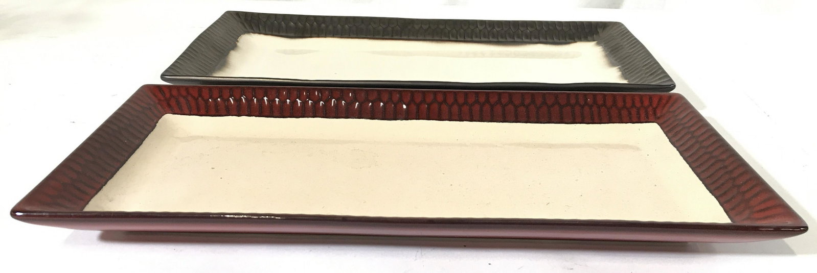 Lot 2 TASTEFULLY SIMPLE Porcelain Serving Dishes: Lot includes 2 rectangular shaped porcelain serving tray. Underside of one is marked TASTEFULLY SIMPLE while the other is marked CRATE & BARREL. One has red toned rim while other has bronze toned rim.