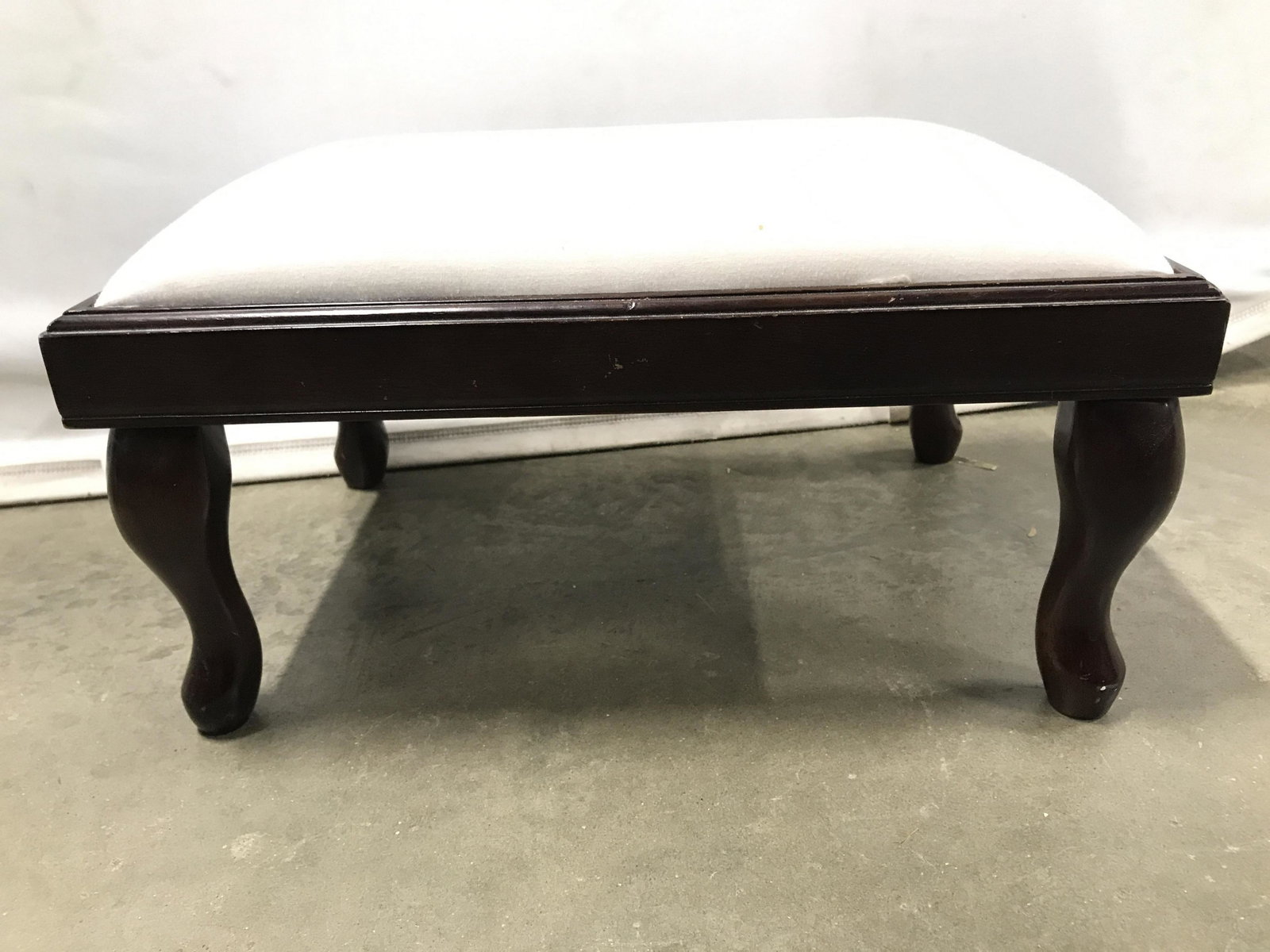 THE BOMBAY COMPANY Upholstered Footstool: Underside of piece is marked THE BOMBAY COMPANY. Footstool is upholstered with white toned fabric. Piece has brown toned carved wooden legs. Piece measures approximately 17 inches long 12 inches wide,