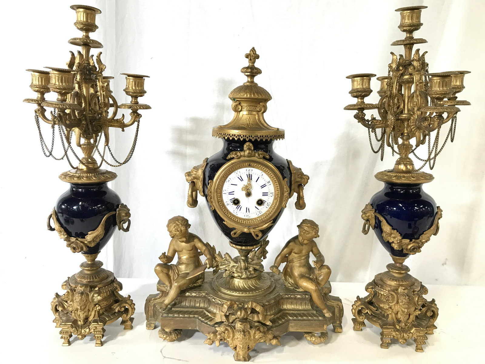Antique 3 Piece Gilded Metal Garniture Set: Lot includes 1 clock and 2 candelabras. Clock has intricate gold toned gilded metal detail along with seated cherubs and lion head detail on sides with swinging loop detail. Finial on top of piece has