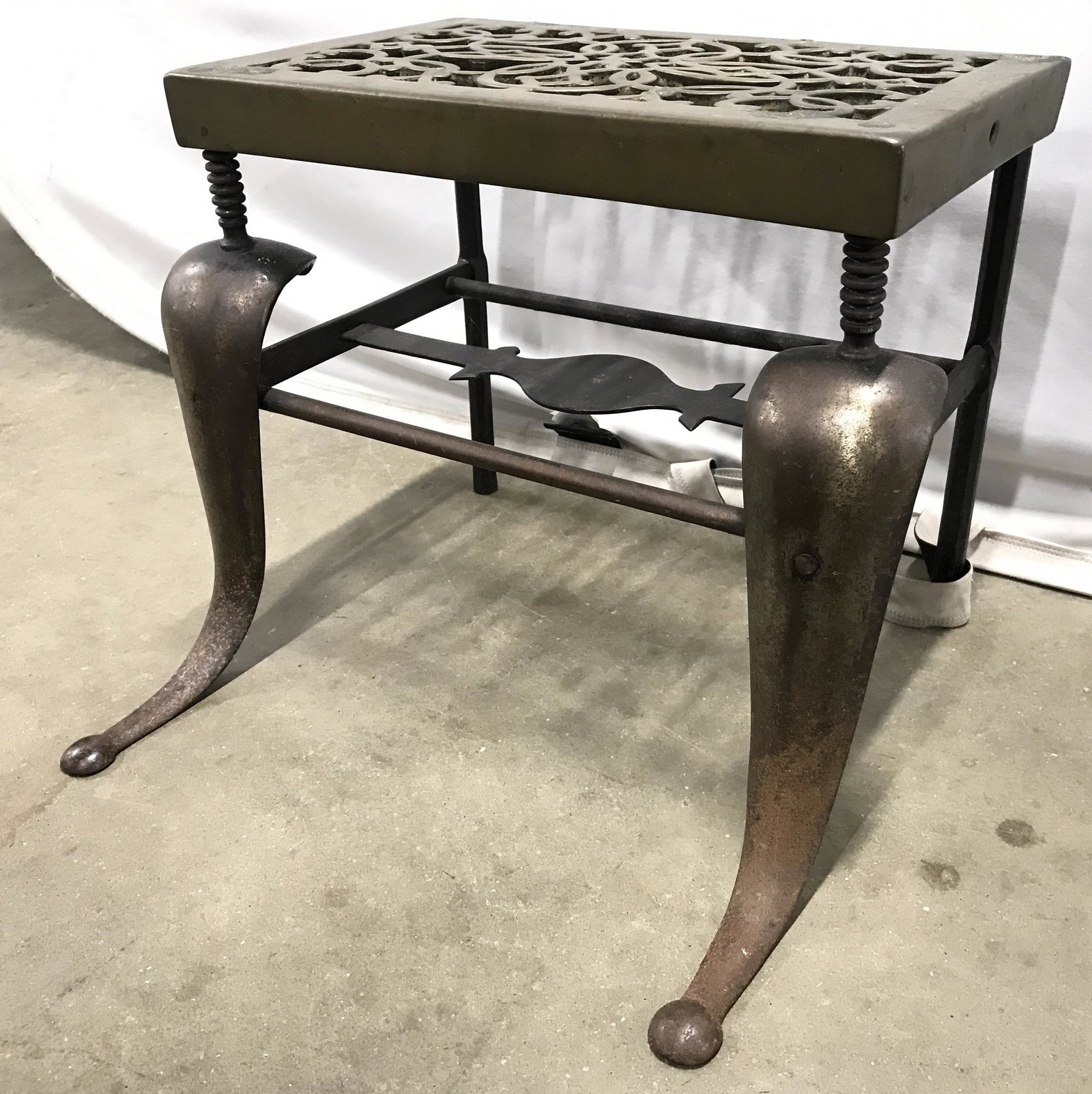 Vintage Brass Fireplace Footman Trivet: Footman trivet is vintage possibly antique. Piece is gold & black toned. Piece is brass in some areas. Contains 2 curved legs with Round structures at feet. Top of piece has pierced detail. Measures a