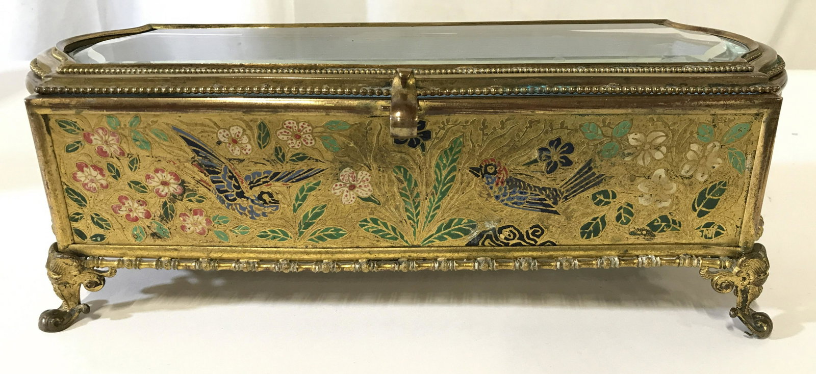 Gold Toned Glass Lid Footed Brass Keepsake Box: Keepsake box is gold toned. Piece appears to be brass. Piece is vintage. Contains multi toned bird, leaf, and floral detail. Piece has gold toned ornate feet. Lid is glass and flips open. Interior has