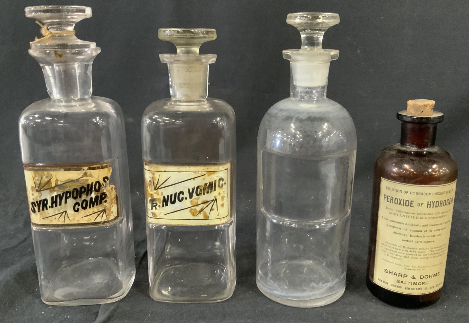 Lot 4 Vintage Apothecary Bottles: Lot 4 Vintage Apothecary Bottles, labels read ‘Syr Hypophos Comp’, ‘TR Nuc Vomic’, ‘Sharp & Dohme Peroxide of Hydrogen’, tallest bottle measures 8 5/8 inches tall x 2 7/8 in diameter, smal