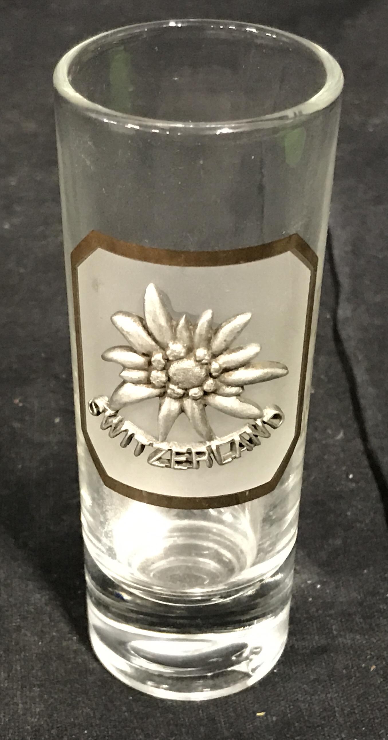 Switzerland Shot Glass: Switzerland shot glass, metal plaque to front shows a flower and reads Switzerland, tall clear glass, measures 4 inches tall, written to the side of the glass reads Geneve, Shot glass, glass, glasswar