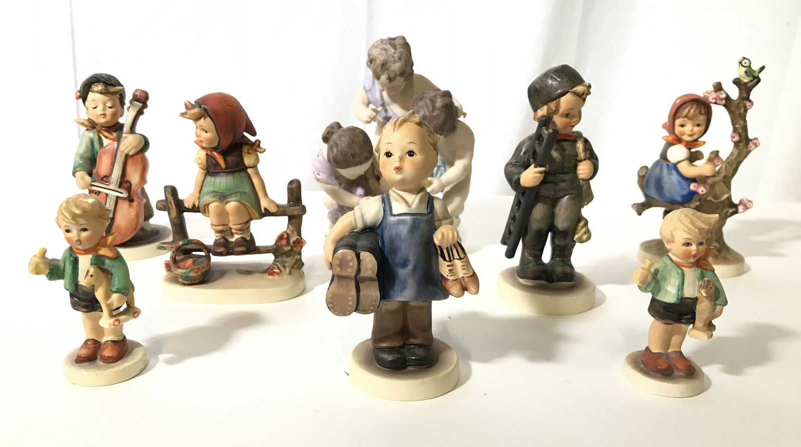 Lot 8 Porcelain HUMMEL Children Figures: Most pieces are marked GOEBEL W GERMANY. Includes 1 child with violin, 1 child sitting on fence, 1 child with ladder, 1 child sitting on branch, 1 grouping of cherubs, 1 child holding shoes & 2 childr