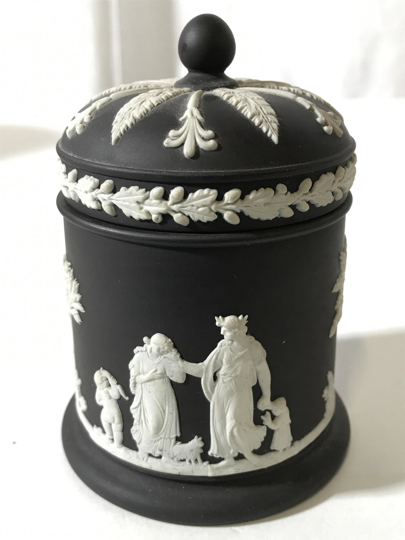 Footed Black & White Toned WEDGWOOD Trinket Box: Underside of piece is marked WEDGWOOD ENGLAND. Piece is black toned with white tree, human Figural and leaf detail. Trinket box includes lid with spherical shaped structure at top. Piece is footed. Me