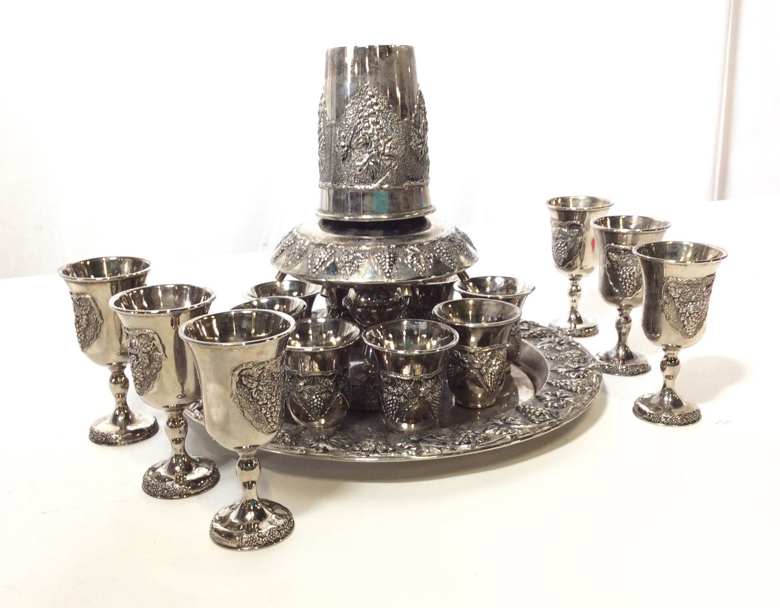 Mid Century KARSHI Repoussé Wine Divider Set: Total pieces 17. Signed KARSHI Repoussé Metal Wine Divider Set, Hebrew Ware, depicts Grape Bunches. Platter with Kiddush Goblets, Wine dispenser positions over ‘ shot glass’ Goblets. Verso Of Pla