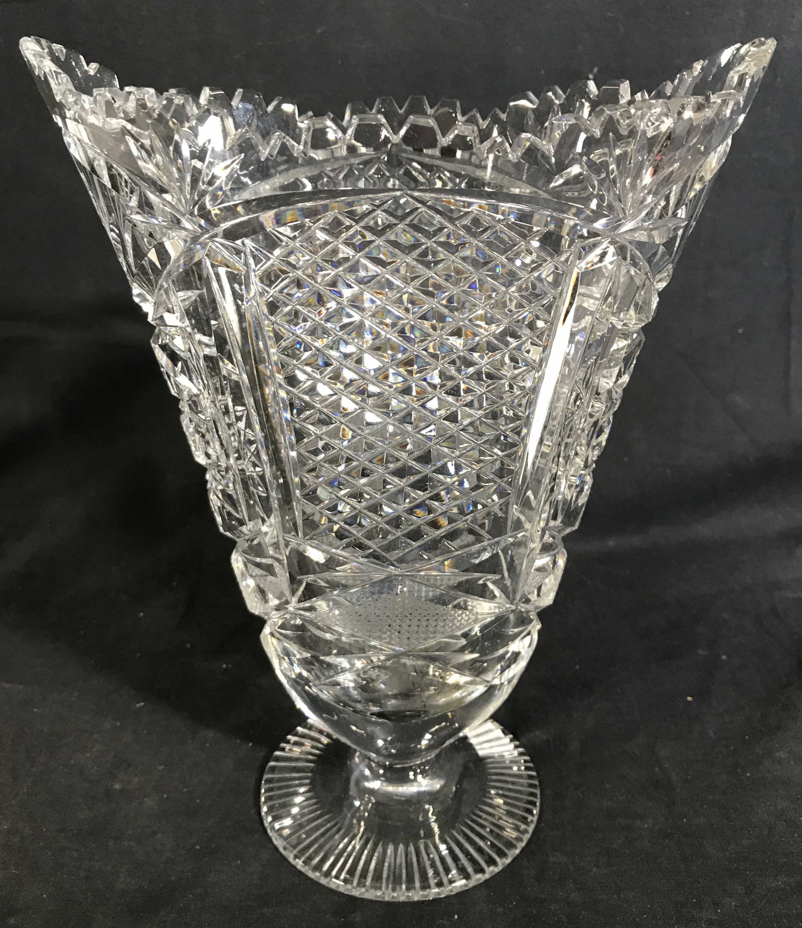 Toothed Cut Crystal Pedestaled Vase: Vase is cut crystal. Piece has intricately cut geometric detail. Rim of vase is toothed. Base of vase is pedestaled. Vase measures approximately 10.5 inches tall 8 inches wide 5 inches deep. Vase, ped