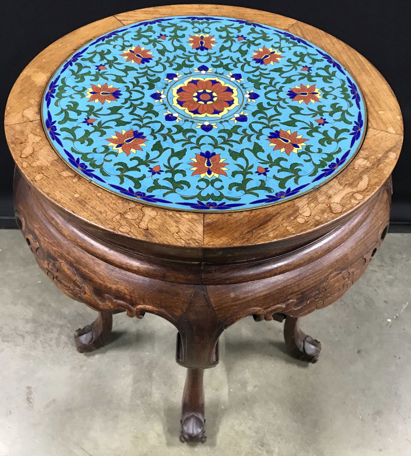 Enamel Top Asian Carved Wooden Side Table: Side table has enameled cloisonné top with intricate multi toned floral, vine, & leaf detail. Table has 5 intricately carved wooden legs. Tabletop has round shape. Piece measures approximately 20 inc
