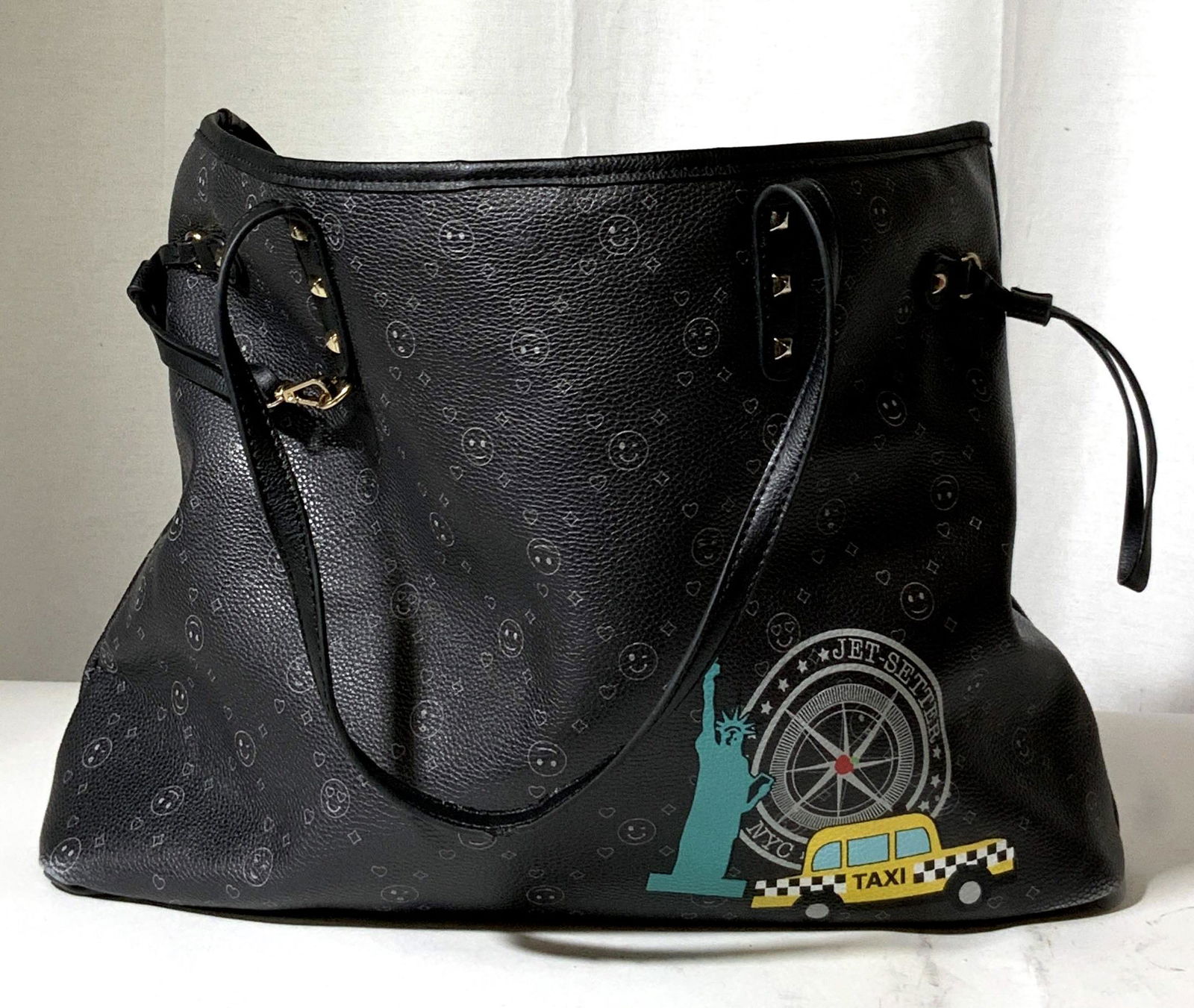 New York Taxi Tote Bag: Black tote bag with emoji designs throughout. Bottom right corner decorated with Statue of Liberty, compass, and taxi. Wording reads: NYC, Jet-Setter. Exterior is possibly patent leather, interior sof