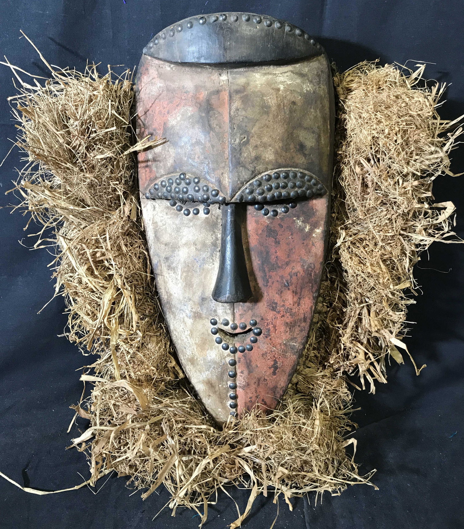 Gabon Mitsogo Mask: Gabon Mitsogo Mask, origins of the Mitsogo people can be found in Gabon Central Africa, painted with white black and red pigment, raffia decorates the side of the mask besides the top border, metal st