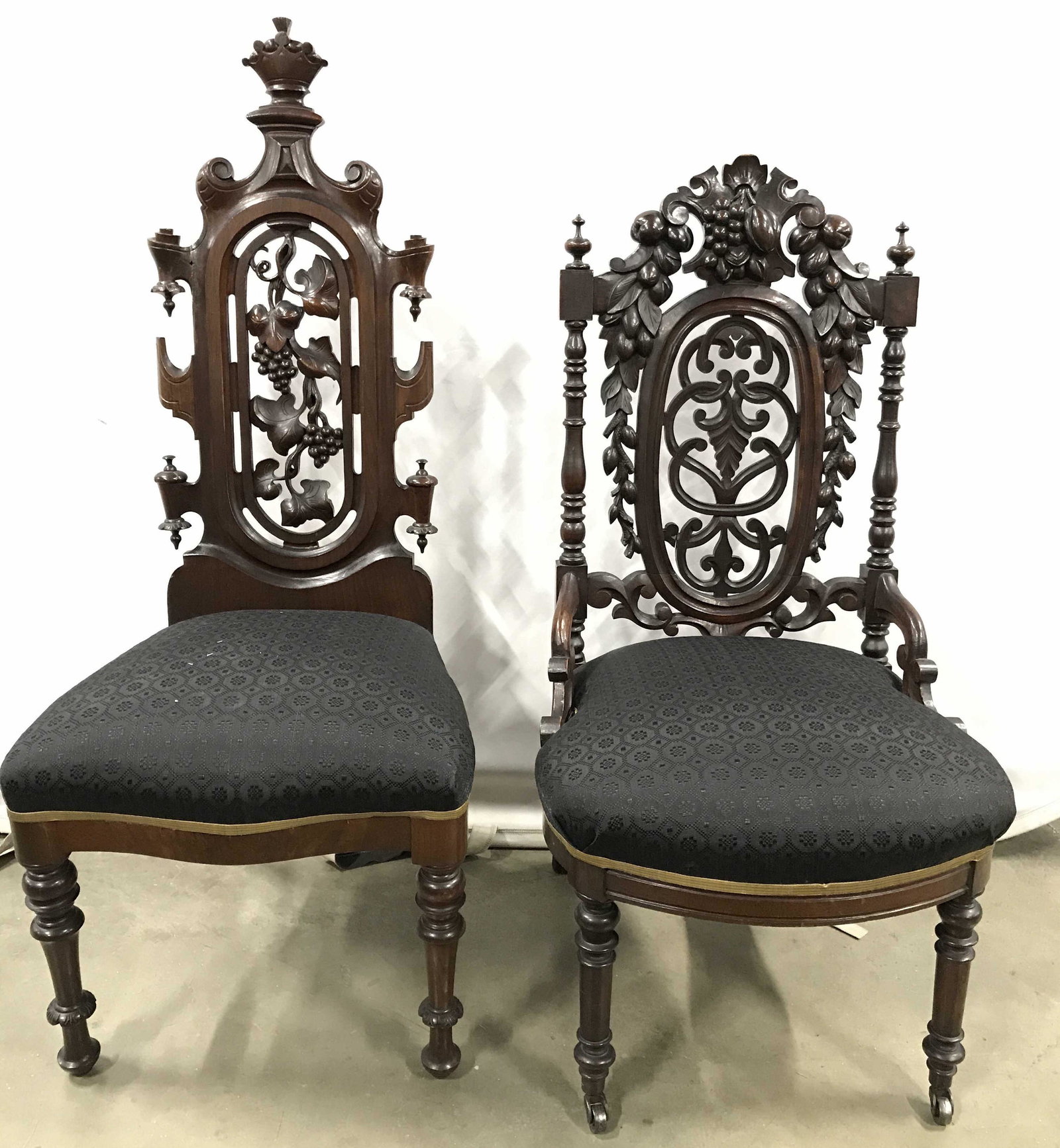 Lot 2 Antique Carved Wooden Him & Hers Chairs: Chairs are vintage appear to be antique. Each have intricately carved wooden grape, vine and leaf detail. Front 2 legs of 1 chair has casters. Top area of other chair has carved crown detail. Him and 