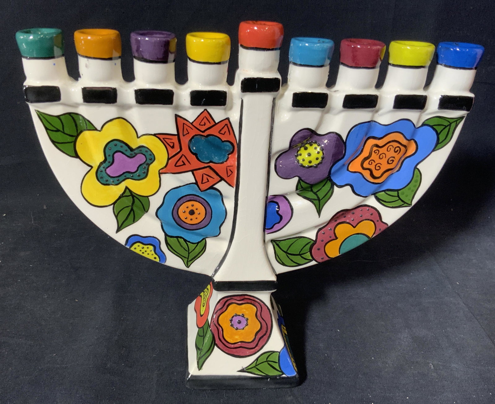 Artist Signed Hand Painted Ceramic Menorah: Paste up dotted decor on bright colorful hand painted flowers, white background. Underside signed JOYCE. Each candle holder with different color tip. Measures 10 in wide, 8.75 in. Tall. Signed Artist,