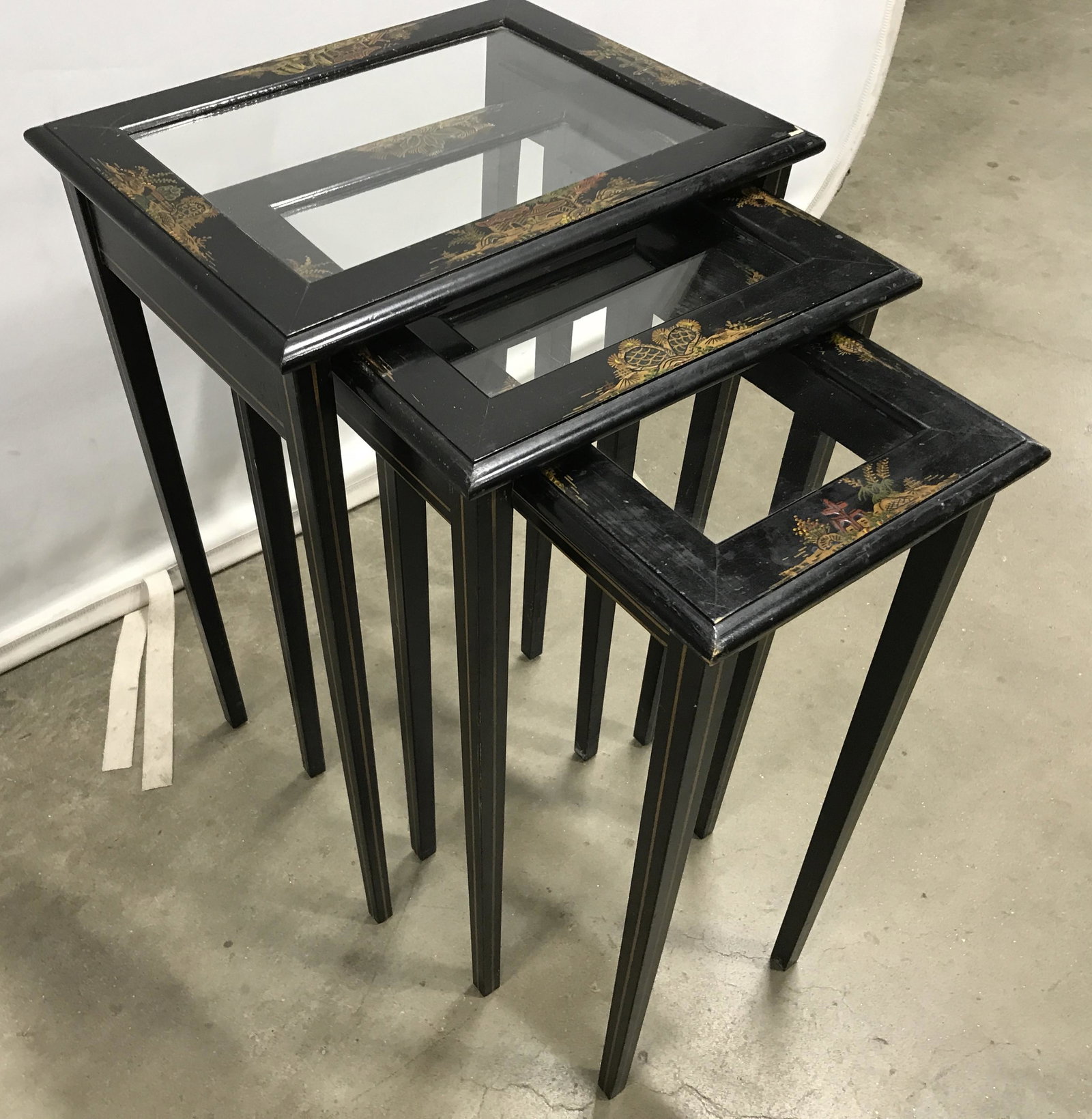Set 3 Vintage Nesting Tables: Set of three nesting tables, crafted from wood, top is glass. Painting depicts various structures and landscapes. Tables are colored black, with gilt paint highlights. Largest piece Measures approx 22