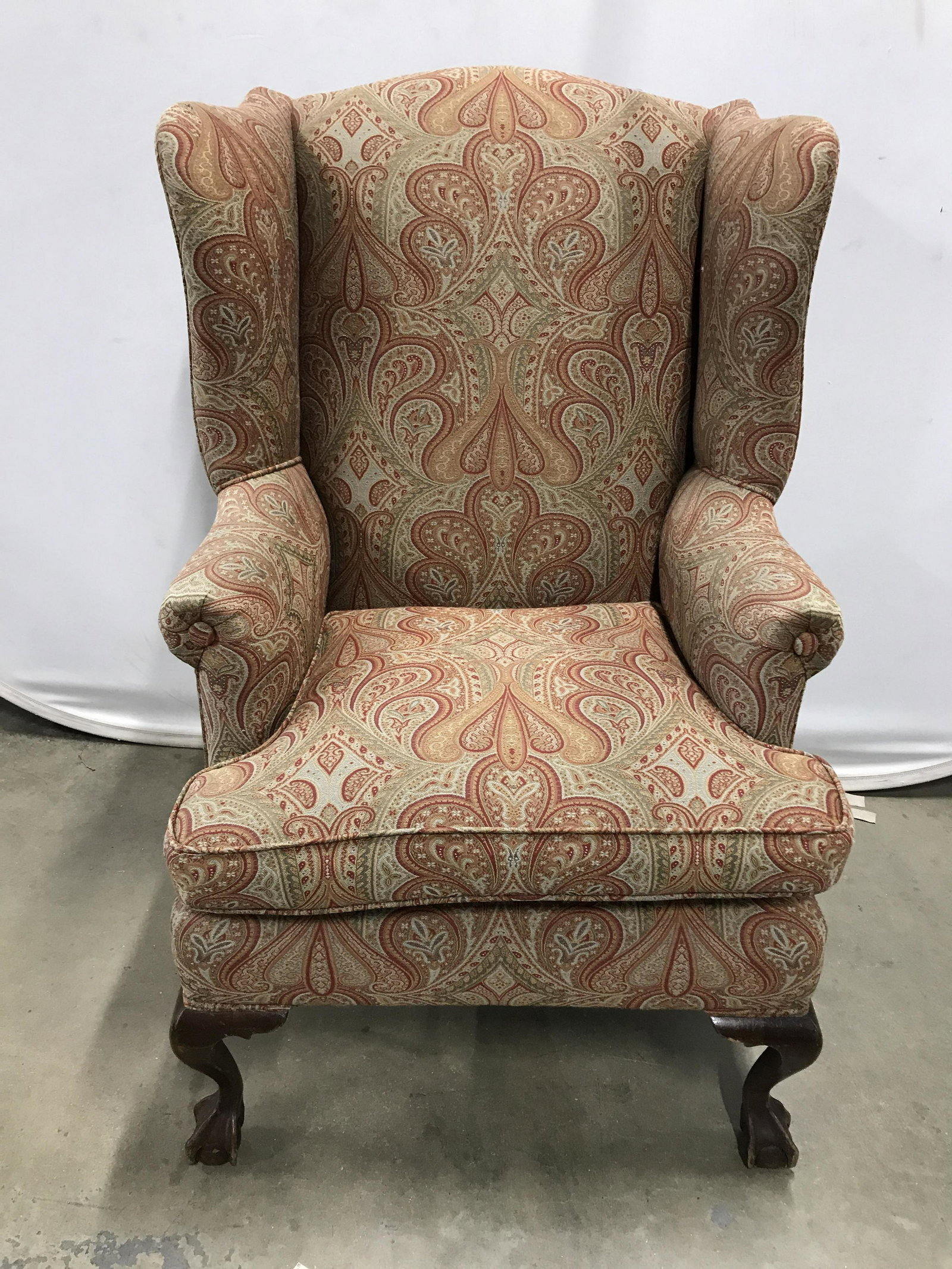 Vintage Paisley Wingback Armchair: Vintage upholstered armchair. Chair is wing backed, front legs are carved and resemble claws grasping spheres. The upholstery is decorated with an all over paisley print. Upholstery secured to frame i