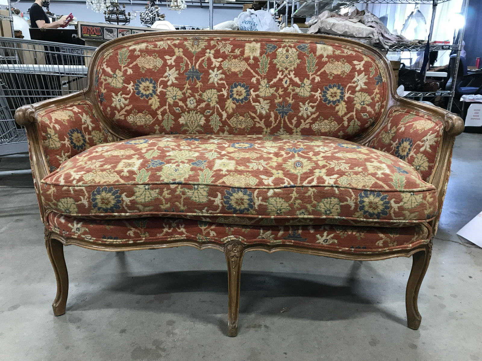 Vintage Carved Wooden Upholstered Loveseat: Loveseat has carved wooden armrests and 5 legs. Piece has multi toned floral leaf and vine detailed upholstery. Top of legs have carved floral detail. Piece measures approximately 53 inches long 36 in