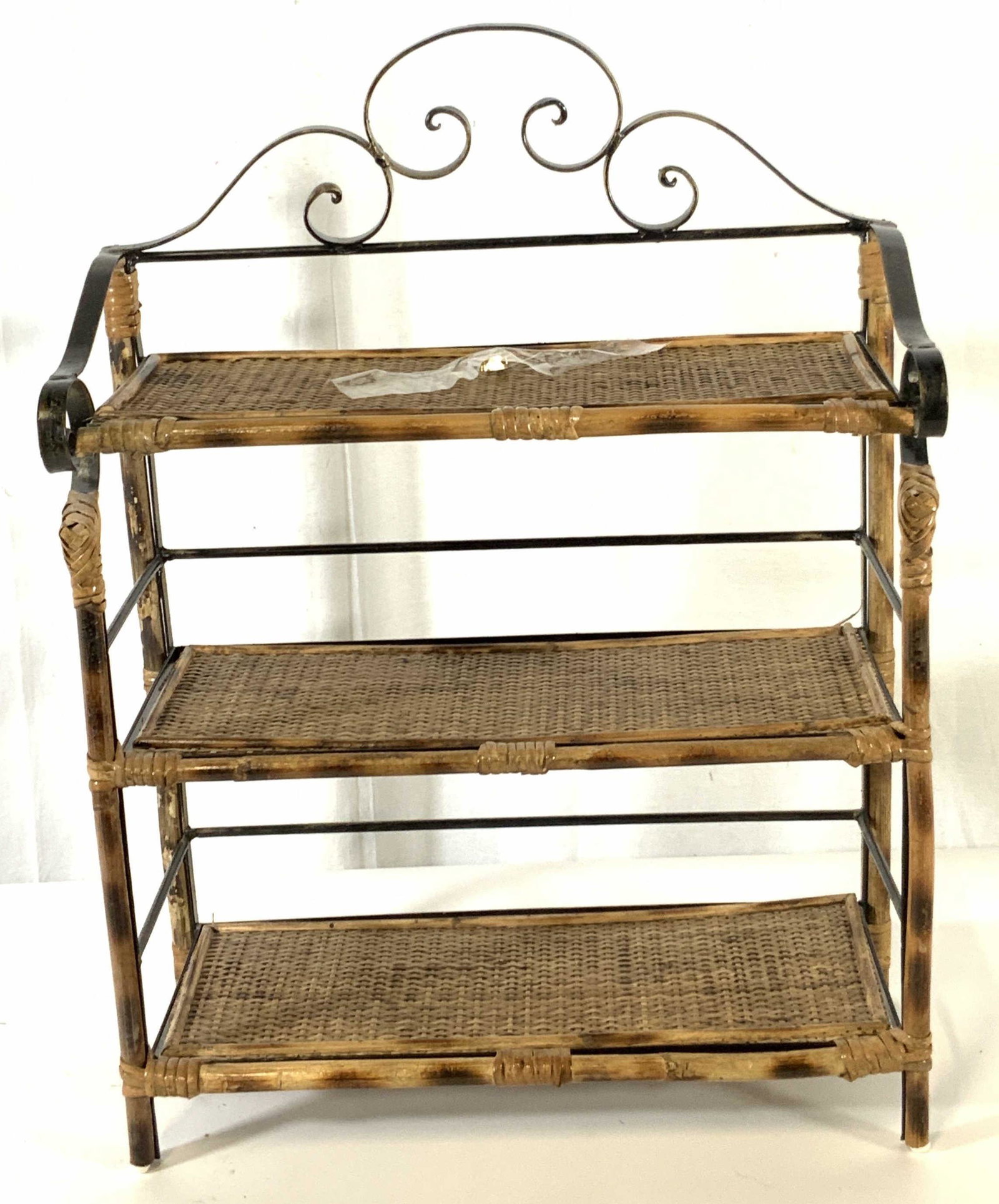Vintage Rattan & Metal Three Tier Floor Shelf: Floor shelf made from metal, wood, and rattan materials. Decorative metal curvature work at the top and sides. Padding on underside of shelf legs. One metal accessory taped to top shelf. 3 shelves for