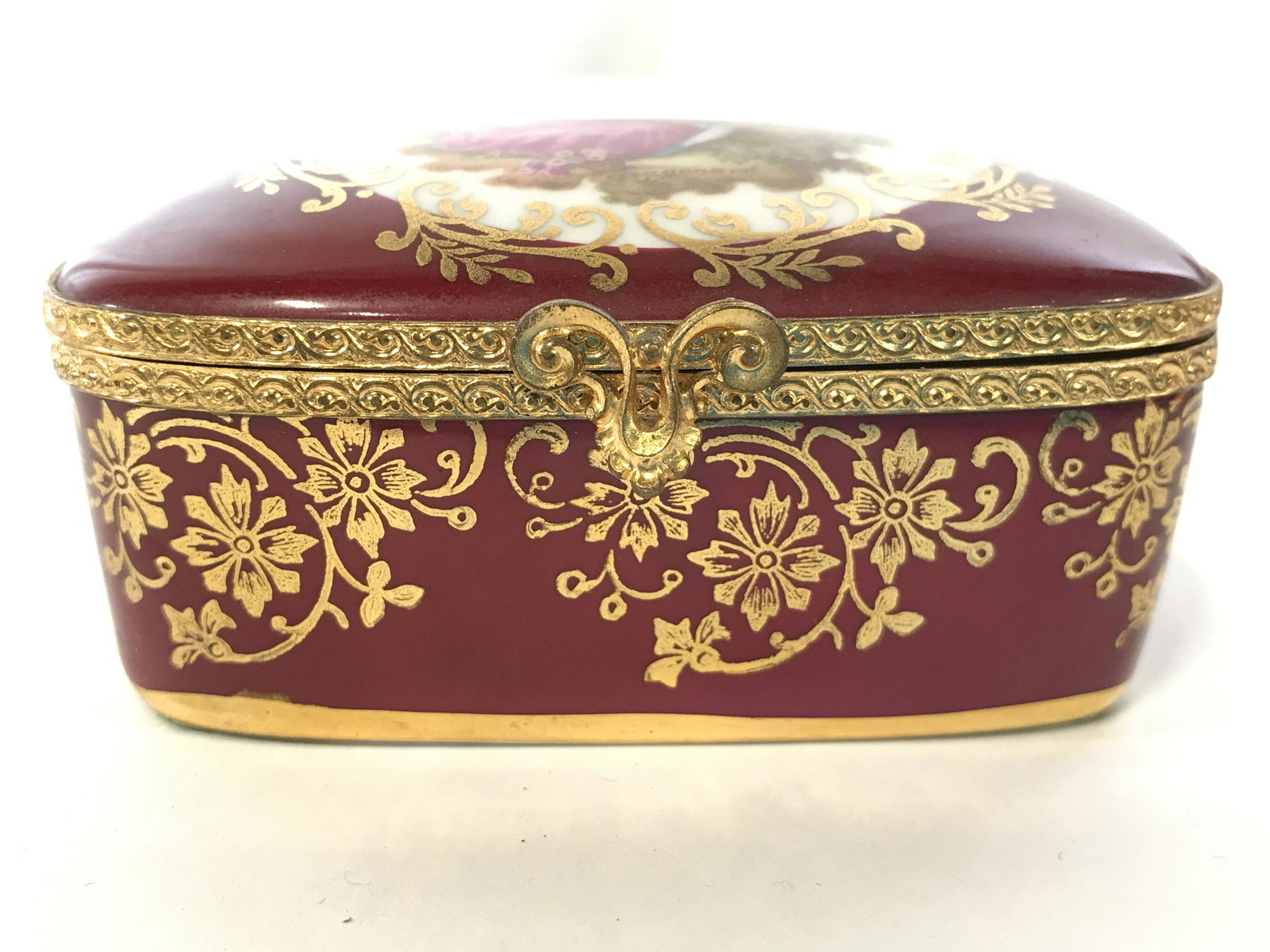 LIMOGES FRANCE Porcelain Trinket Box: Underside of piece is marked LIMOGES FRANCE. Piece is cranberry toned with intricate gold toned ornate detail. Lid has depiction of of seated male & female figure. Piece measures approximately 3.5 inc