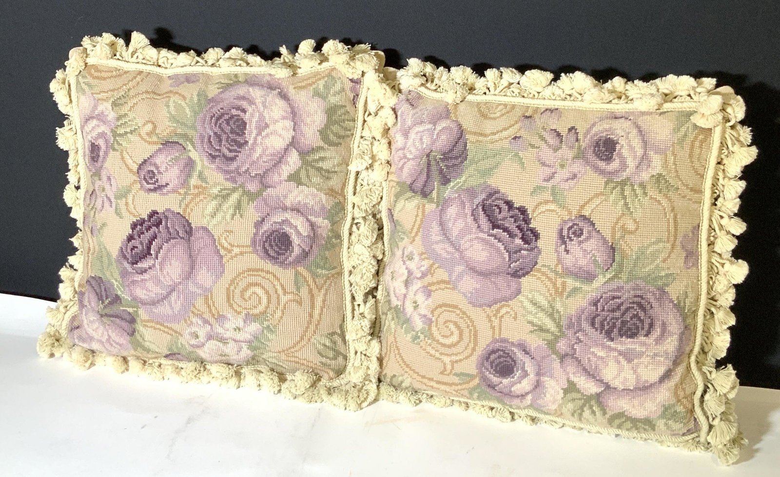 Pair Floral Needlepoint Throw Pillows: 2 Needlepoint throw pillows with purple flower designs. Muted earth toned colors. Tasseled trimming along all edges. Underside soft textile. Each pillow has a zipper. Approx. 18 x 18 in. Sofa accessor