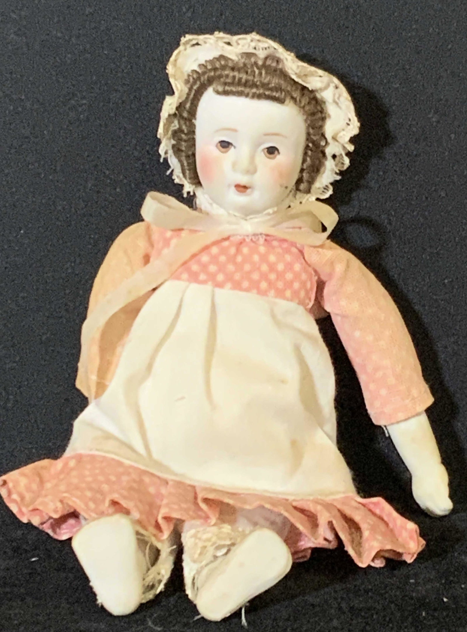 Vintage Hand Painted Porcelain Doll: Vintage, possibly antique, doll with hand painted porcelain face. Brown curled hair, rosy cheeks, and brown eyes. Wearing a pink and white dress & a lacy white bonnet, some discoloration. Approx. 3.5 