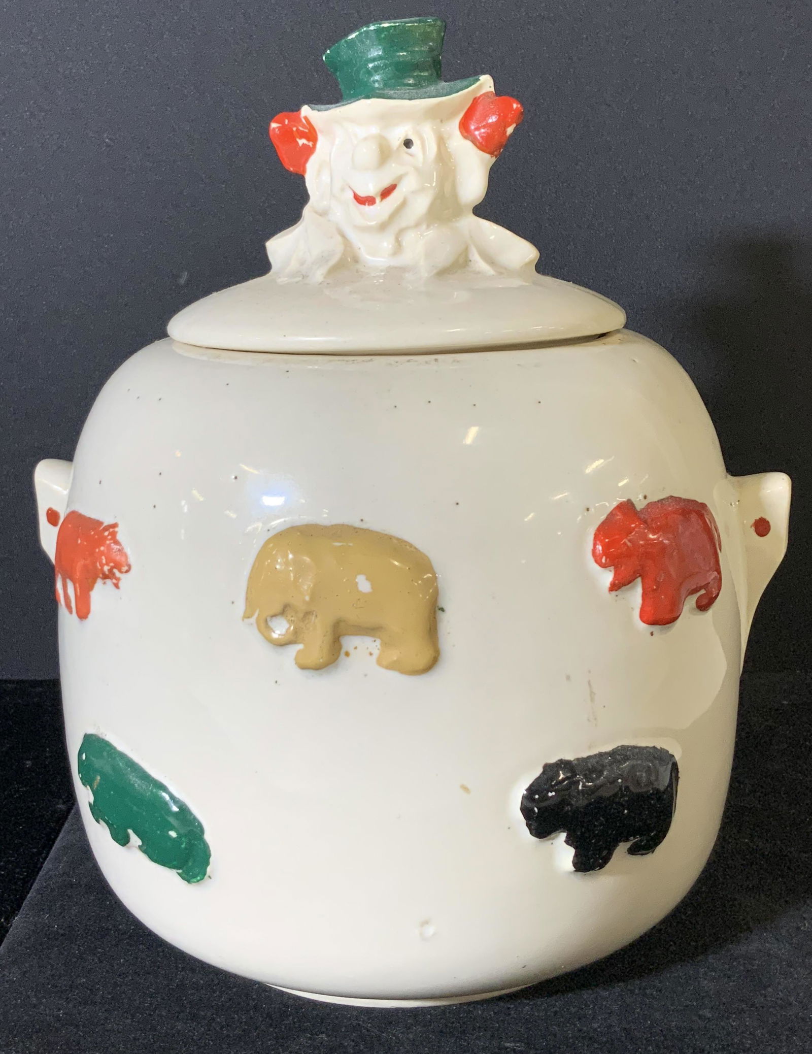 MCCOY Antique Collectible Clown Cookie Jar, Lid: Lidded ceramic cookie jar with clown head figural atop the lid. Clown head is winking, has red hair, and green hat. Handles decorated with one singular red dot on each side. Cookie jar is decorated wi