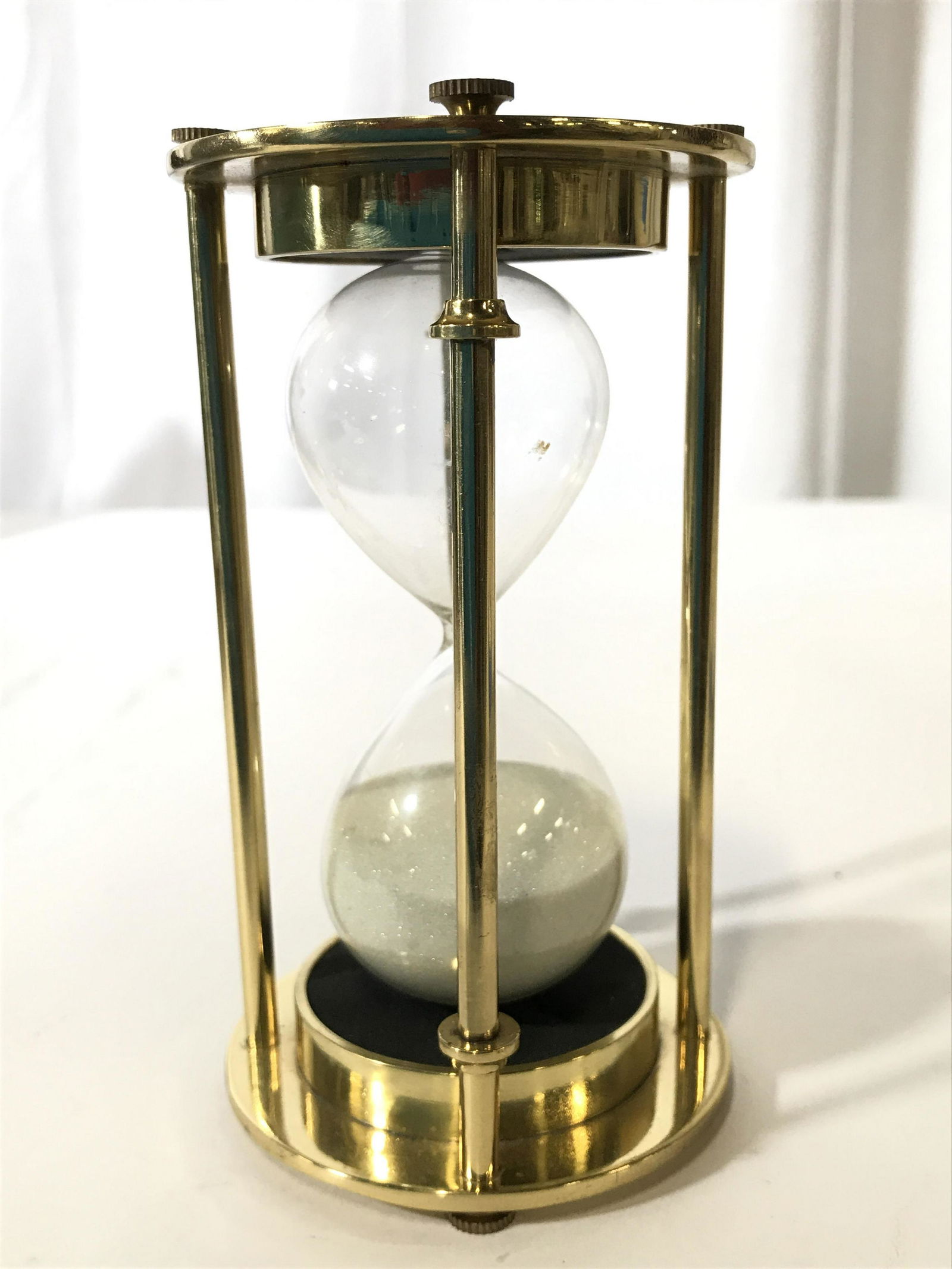 Gold Toned Brass Hourglass: Hourglass is gold toned and appears to be brass. Piece has 4 pillar structures. Top and base of piece are circular shaped. Piece measures approximately 5 inches tall 3 inches wide. desk top accessory,