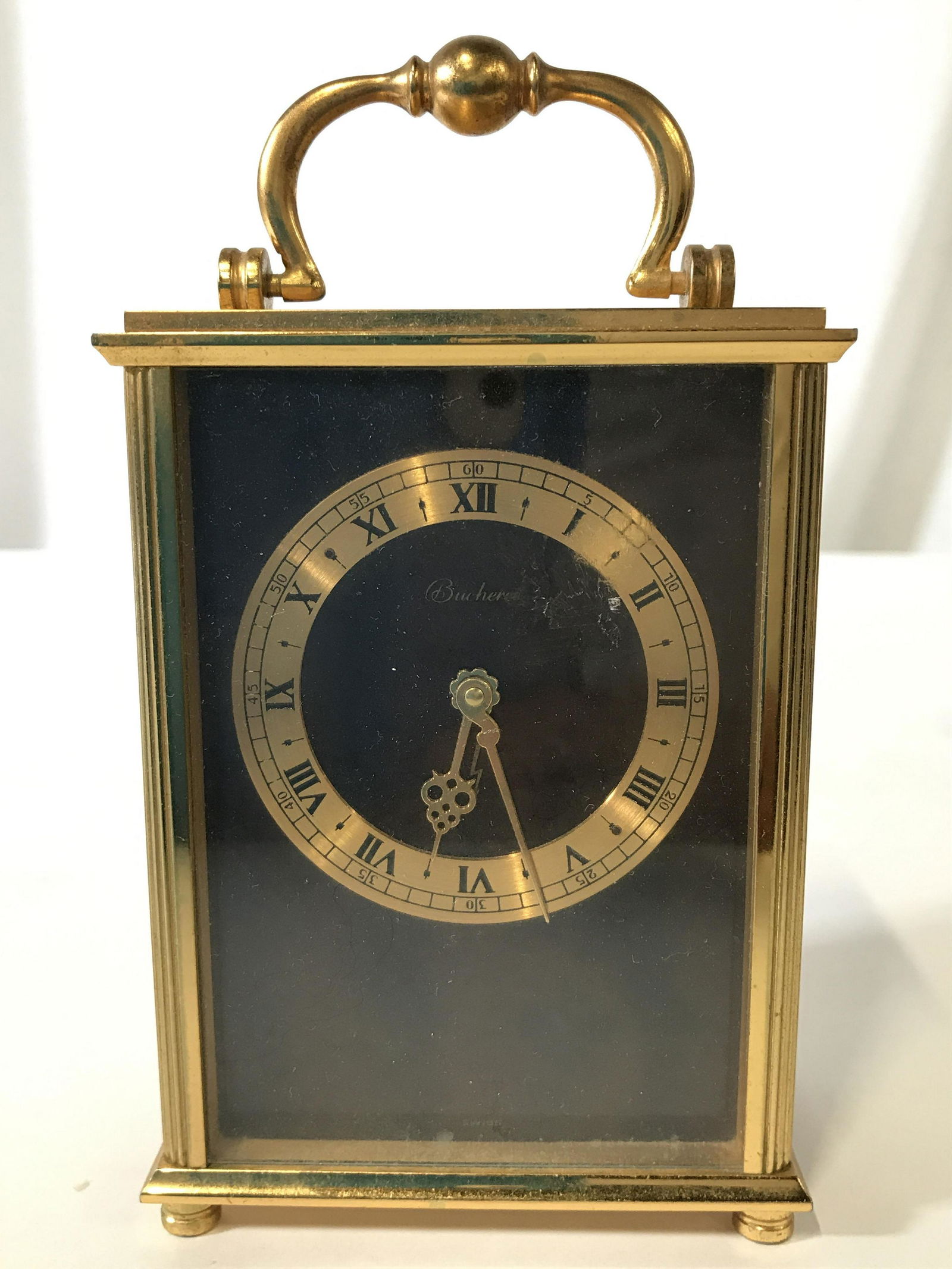 Gold Toned Brass BUCHERER Desk Clock: Clock face reads BUCHERER underside reads SWISS. Clock is gold toned and appears to be brass. Top of piece has handle. Clock face has black toned Roman numerals. Piece measures approximately 6 inches