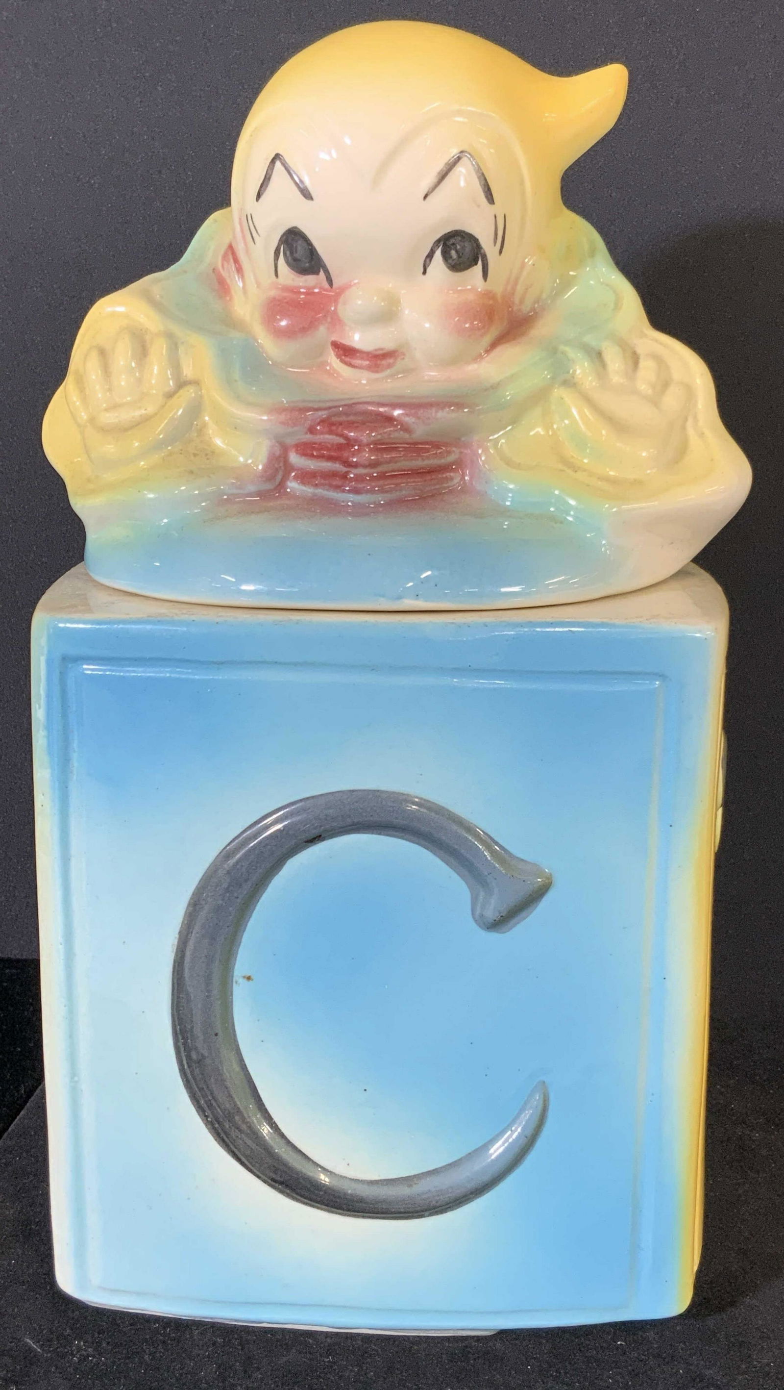 AMERICAN BISQUE Jack In The Box Cookie Jar c40s: Circa 1940s Vintage lidded cookie jar. Depicts a jack popping out of a blue & yellow box. Box is the base of the jar. Letter ‘ C ‘ and the word ‘ cookies ‘ in relief. Lid depicts jack in red,