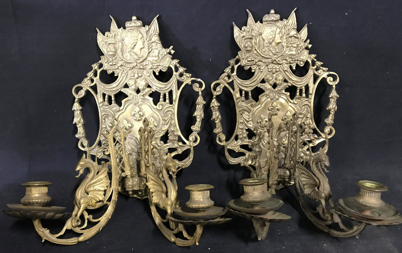Pair Antique Brass Candle Sconces: Ornate candle sconces, pair is antique. Each arm resembles a wyvern. The plaques depict cherubs, flowers, fleur de lis, and a crowned figure. Each measures approx 12.5 inches H, and 13 inches W. Wear 
