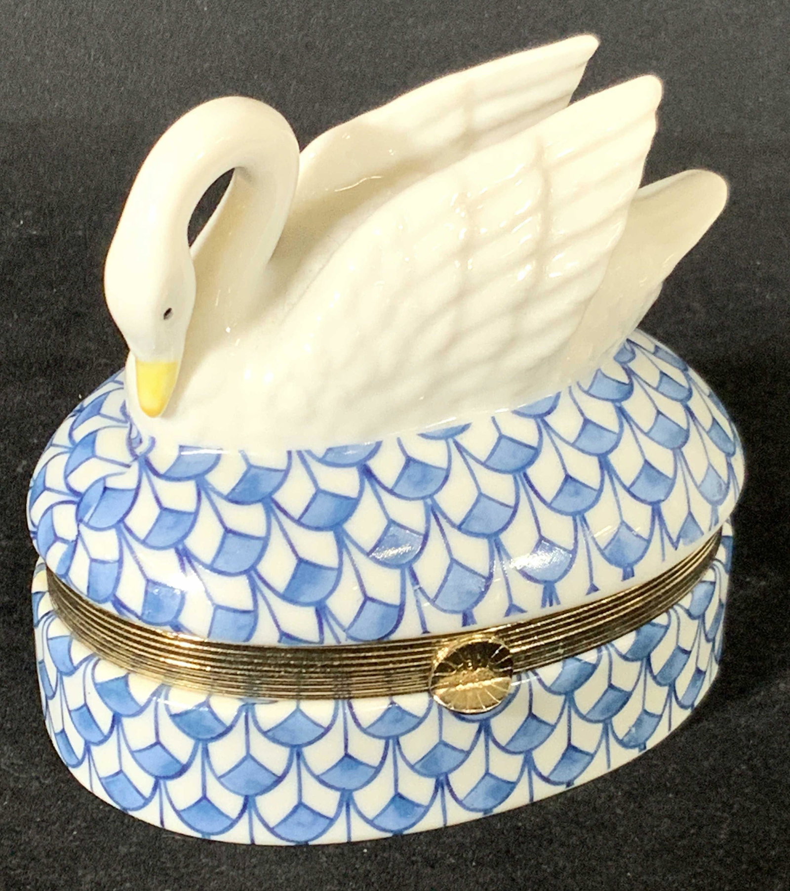 Musical Swan Figural Porcelain Trinket Box: A blue and white patterned trinket box with a hinged lid. Hinge hardware is polished gold tone. Atop the porcelain trinket box is a swan. The swan has embossed feathers. Wings are raised and their hea