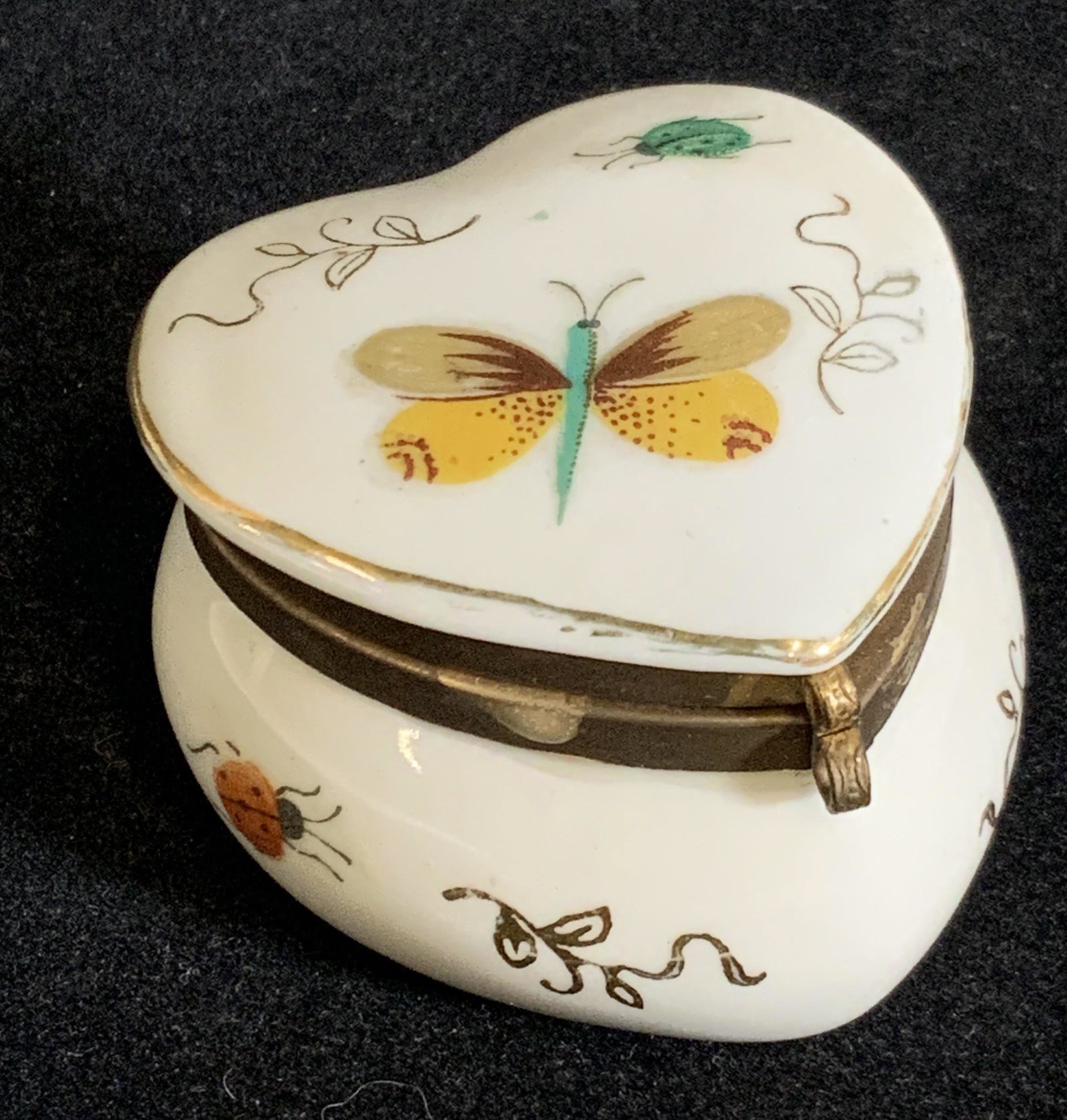 Heart Shaped Insect Motif Porcelain Pill Box: Porcelain heat shaped pill box with hand painted insects and gilt line work. Insects are ladybugs, beetles, and a butterfly. Metal hinge hardware for opening and closing the box. Interior of the pill 