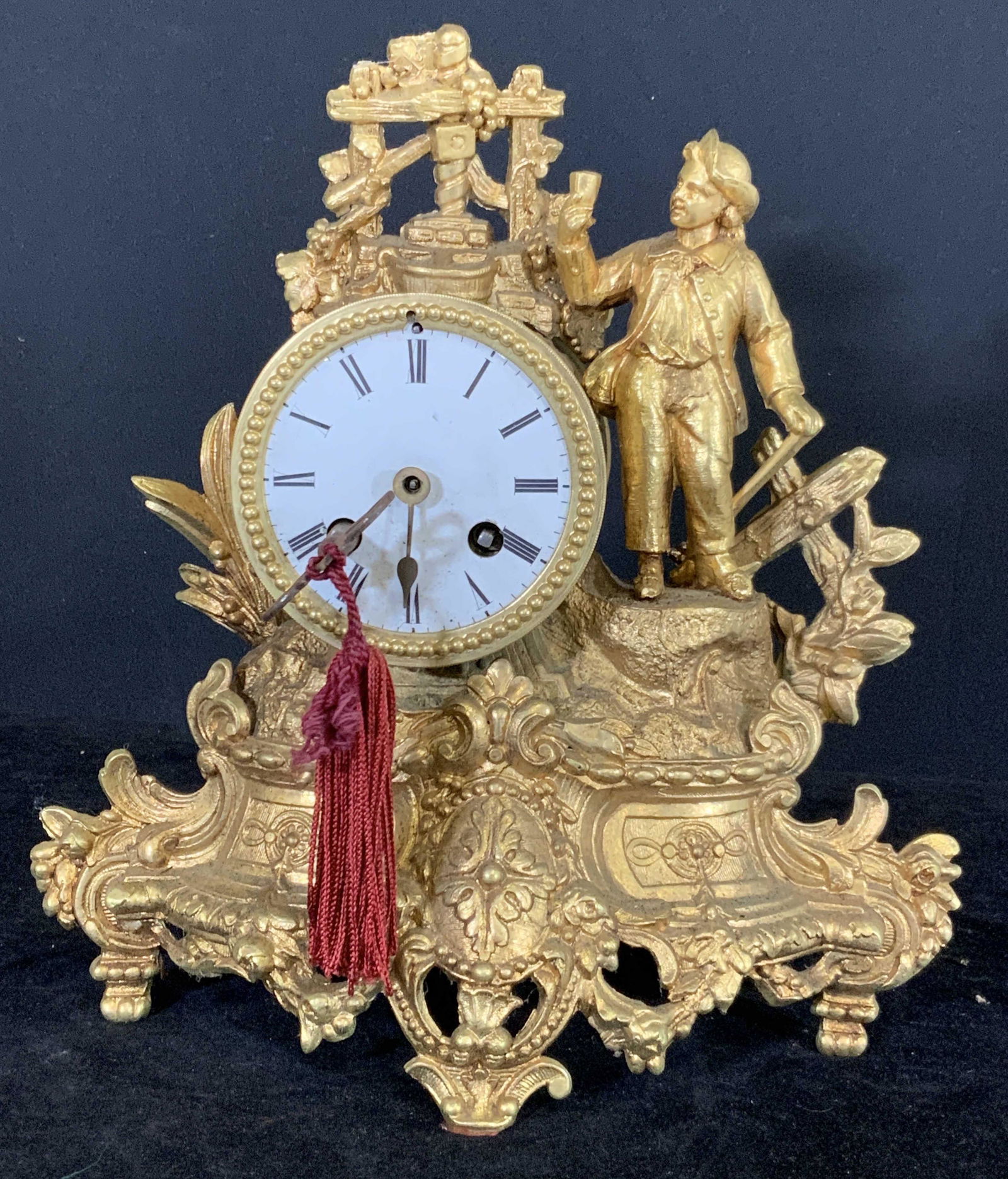 Vintage Gilded Bronze Mantle Clock: Vintage, possibly antique, rococo gilded bronze mantle clock. Decorative feet. Male Figural holding a cup, beside him is a cross. Figural is loose. A key w/ red tassel hangs from clock face. Clock unt
