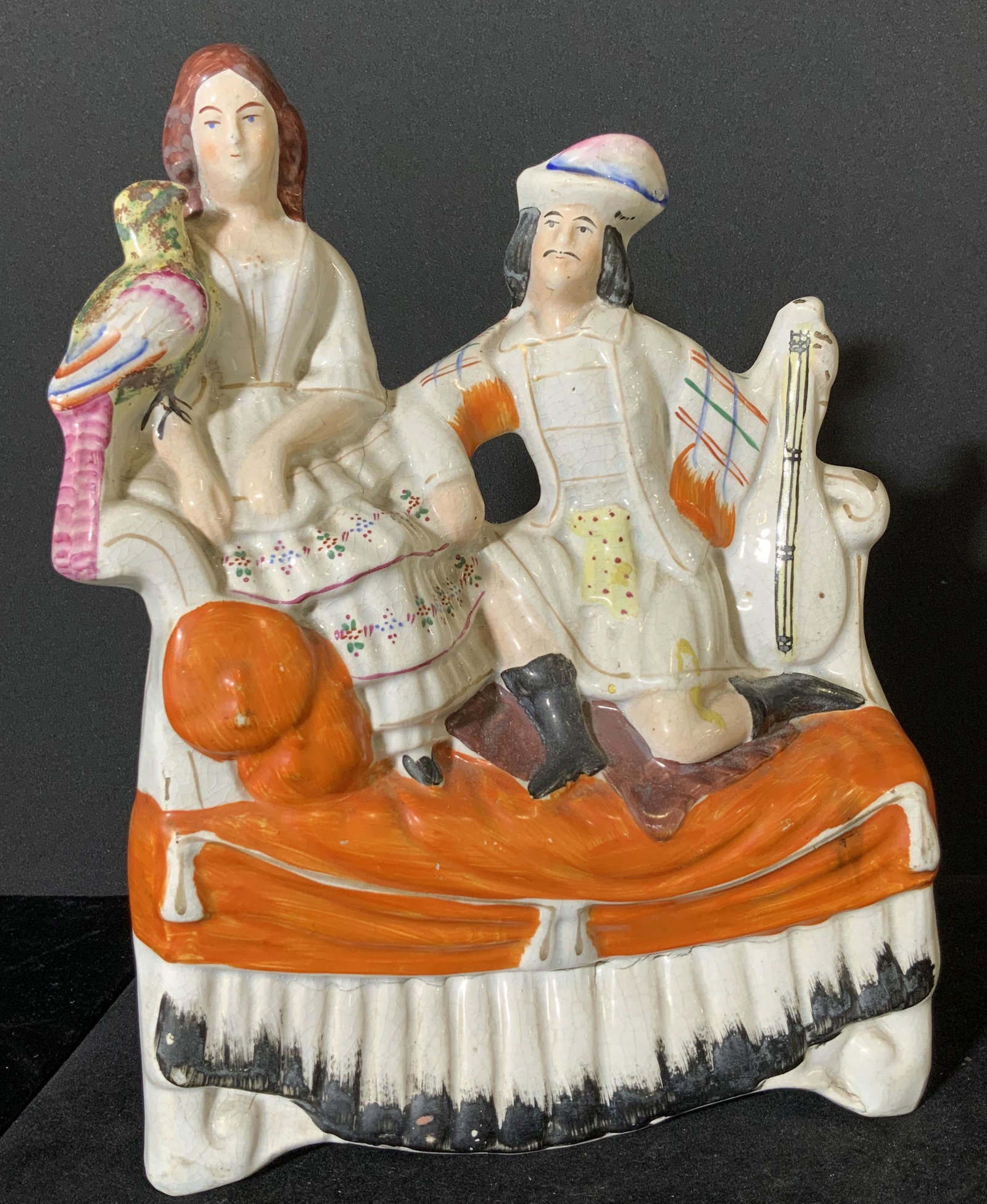 STAFFORDSHIRE Antique Couple On Sofa Figural: Victorian C. 1860 Staffordshire Figural depicting man, woman, and parrot sitting on an elaborate sofa. The man holds a stringed instrument, possibly a mandolin, and the parrot rests on the woman’s a