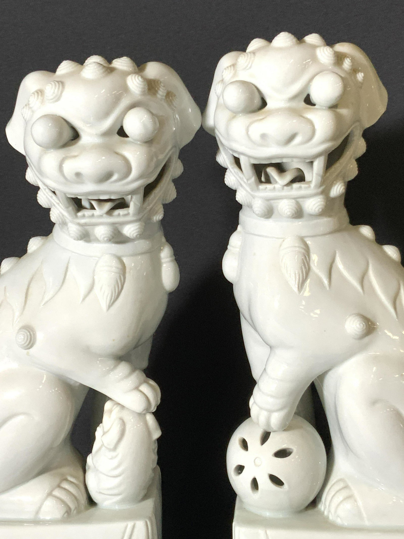 Pair Blanc de Chin Asian Foo Dog Statues: Two white porcelain fu dog / foo dog / temple lion statues. The foo dogs are seated atop white square porcelain stands. They each have their front paw resting on an item. One foo dog has their paw on 