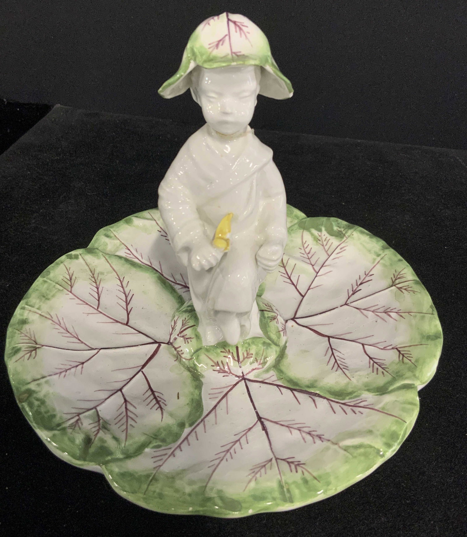 MOTTAHEDEH Sig. Porcelain Child Figural Dish: A porcelain trinket dish with a child Figural holding a yellow flower standing in the center. The dish is hand painted green & maroon, resembles a leaf. The child wears a hat with the same coloring. U