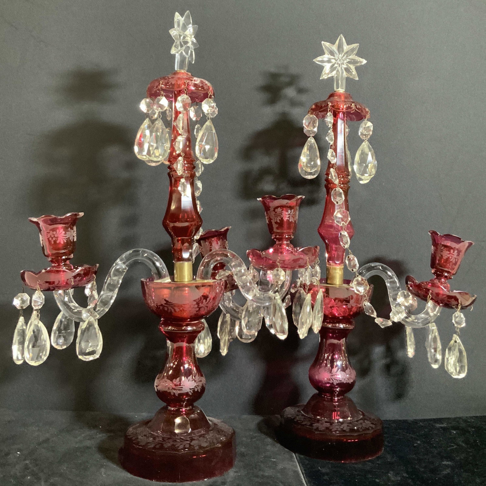 Pair Cranberry Art Glass Tabletop Candelabras: Two cranberry glass candelabras with hanging glass prisms. Some hanging glass is teardrop shaped, others are diamond shaped. At the top there is a star shaped glass accent. On the cranberry glass ther
