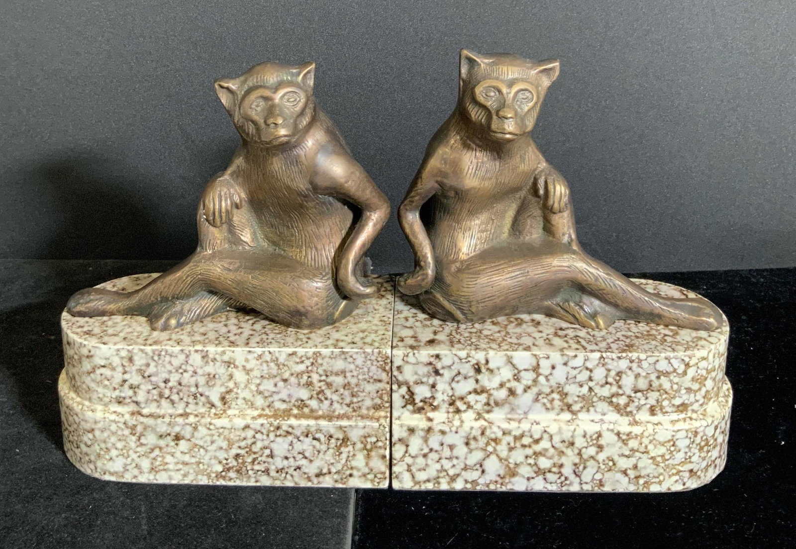 Pair Etched Metal Monkey Figural Bookends: Pair of bookends featuring etched metal monkeys sitting atop marble print composite base. The monkeys are both seated with one arm resting on either knee. The marble print base underside has soft blac