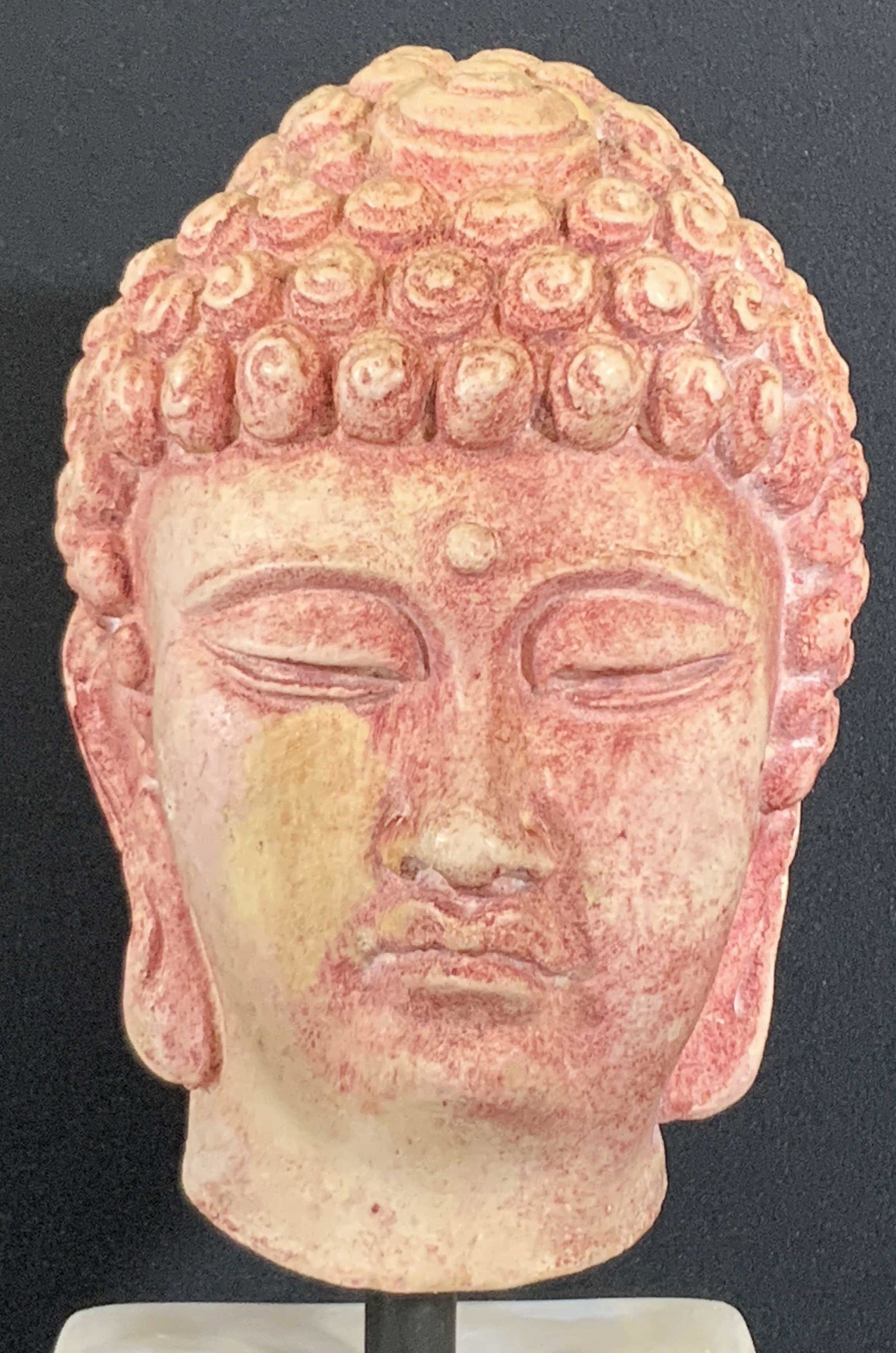 Tabletop Composite Mounted Buddha Bust: A composite Buddha head mounted on metal hardware that is connected to a white composite block. Buddha’s head has light red & white coloring. Signature meditative expression, eyes closed & calm. App