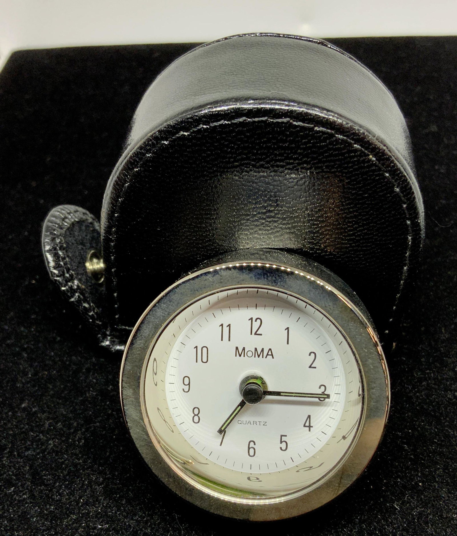 MOMA PORTABLE Quartz Alarm Clock in Case, Hong KG: Signed MOMA silver toned alarm clock, 1 5/8 in round, with flattened underside for upright position. Runs on LR44 battery. Dial to change clock hands. On/ switch for alarm. Verso reads Made in Hong Ko